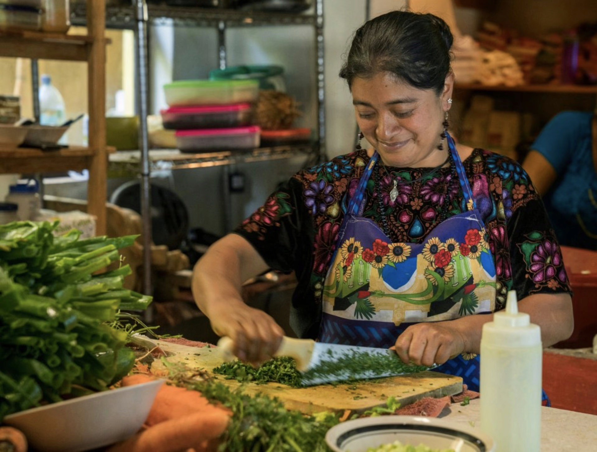 A woman in traditional attire chopping fresh herbs in a kitchen.
