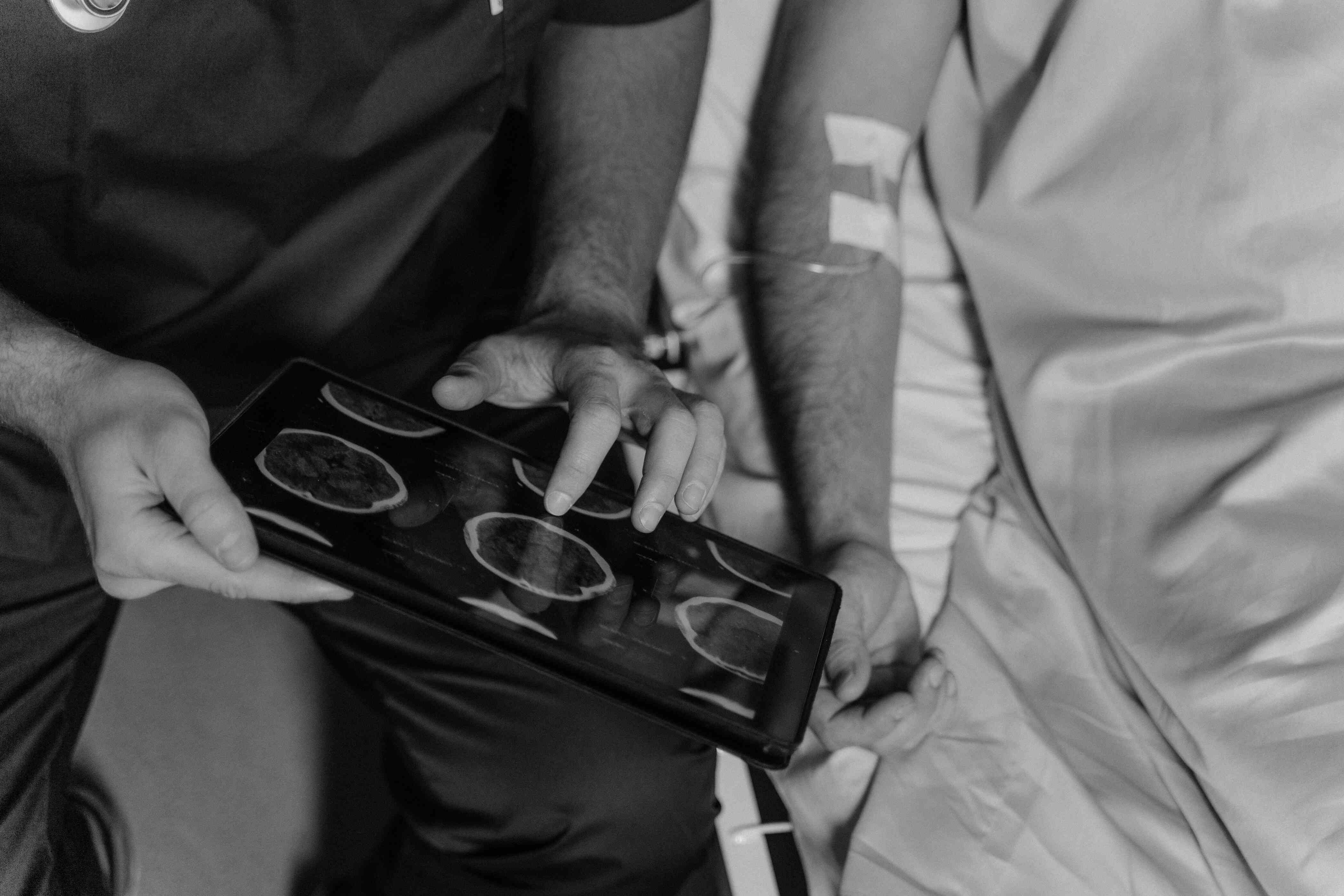 A healthcare professional in scrubs reviewing brain scan images on a tablet while sitting beside a patient in a hospital bed