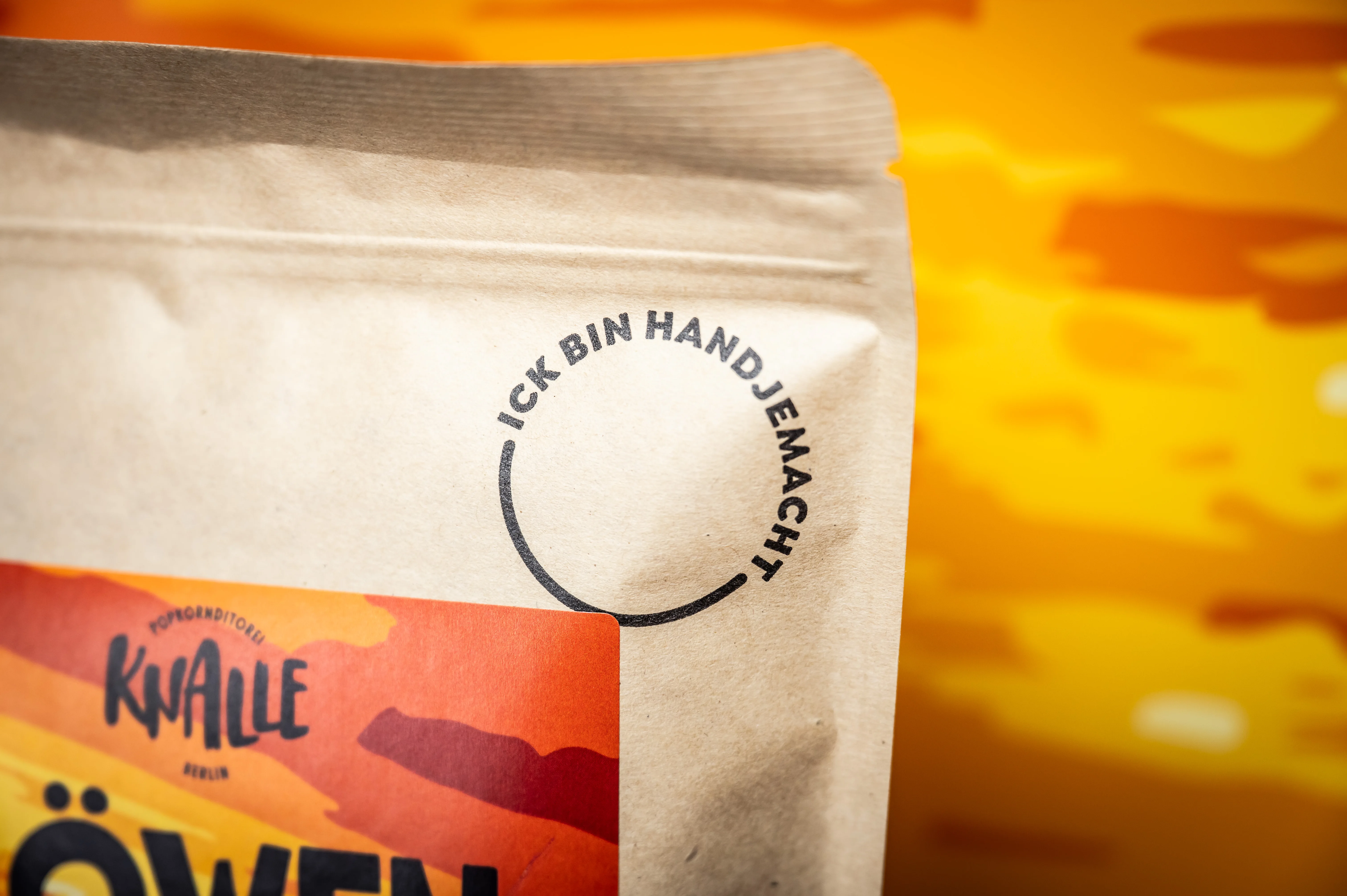 Close-up of a kraft paper KNALLE popcorn bag with the circular stamp Ick bin handjemacht in front of an orange savanna backdrop.