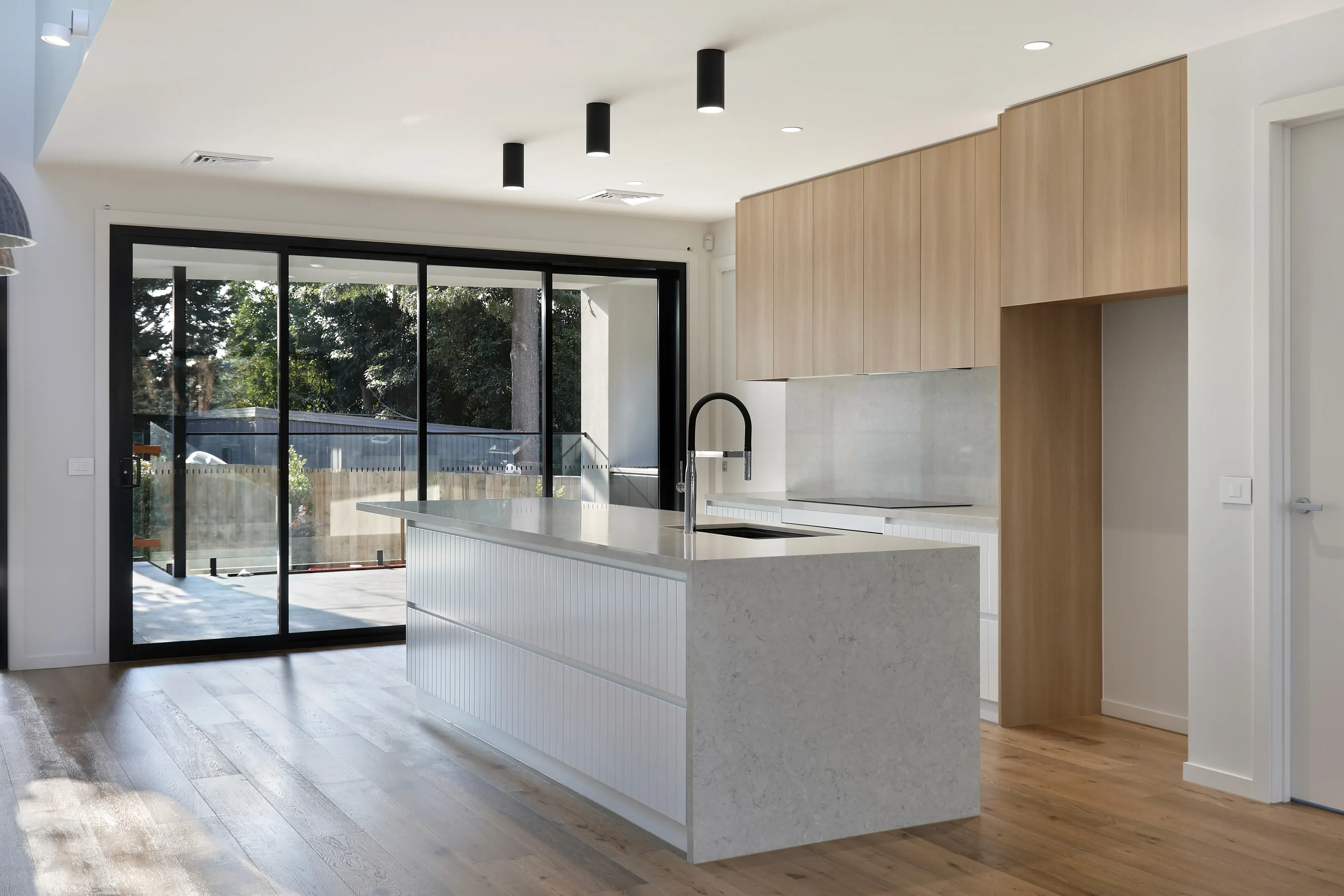 Kitchen with pale-oak cabinetry, a fluted white island and grey stone benchtop, black gooseneck tap, induction cooktop and black sliding doors opening to a deck among the trees