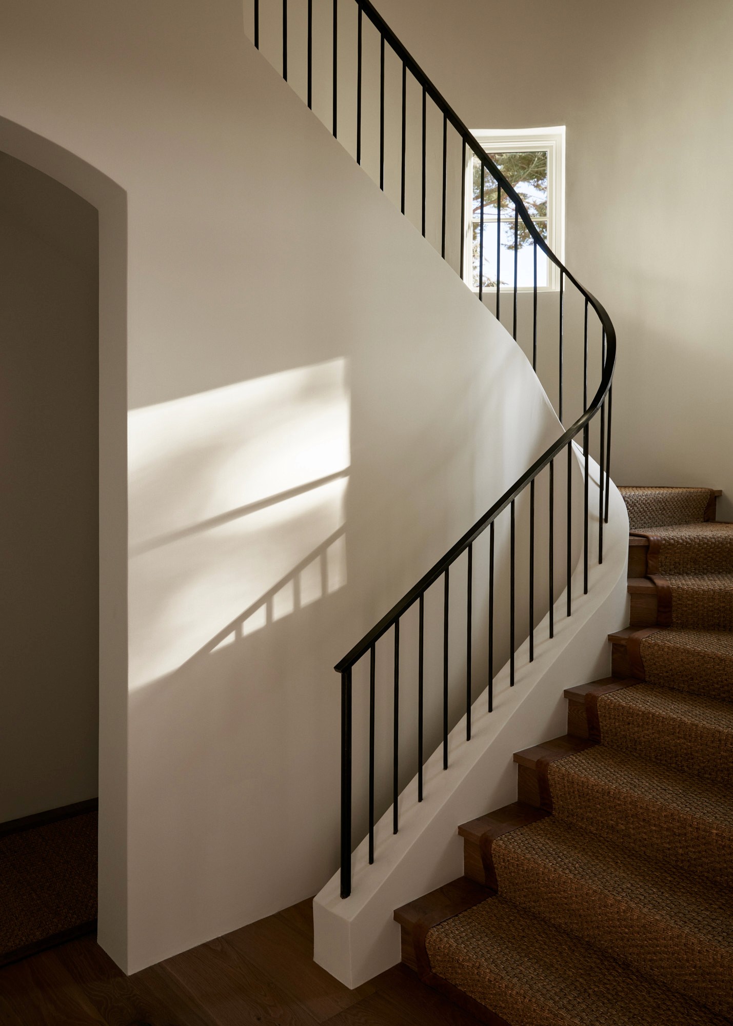 A curved staircase with a hand-forged iron railing and natural fiber runner on oak treads. Sunlight falls across the plaster walls from a small window on the landing above.