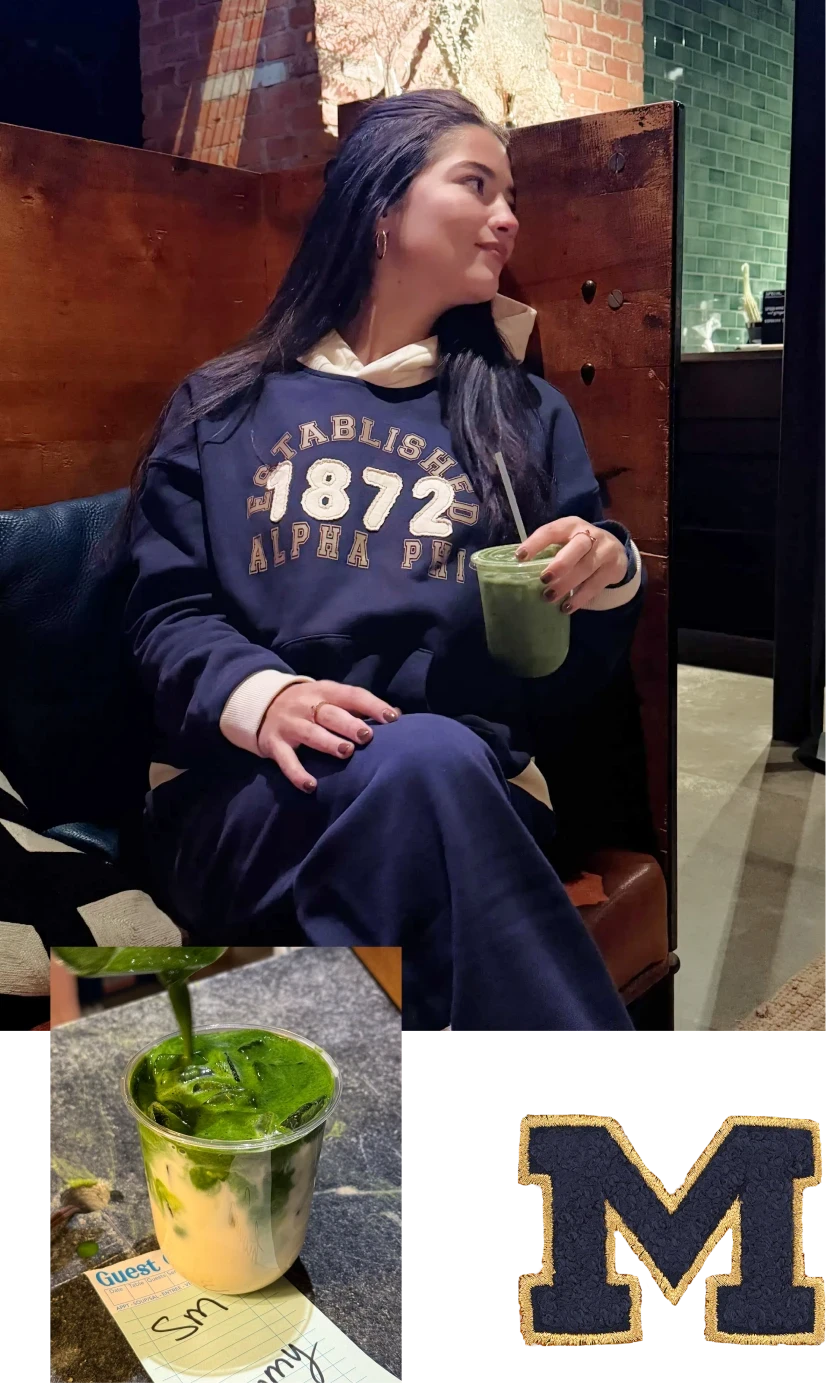 Person seated in a café holding a matcha drink, wearing a navy “Established 1872 Alpha Phi” hoodie