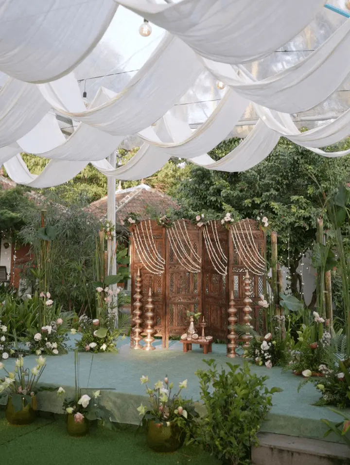 Outdoor wedding ceremony setup at Vedic Village Alleppey with carved wooden screen, white jasmine garland drapes, brass oil lamps and lush tropical garden canopy