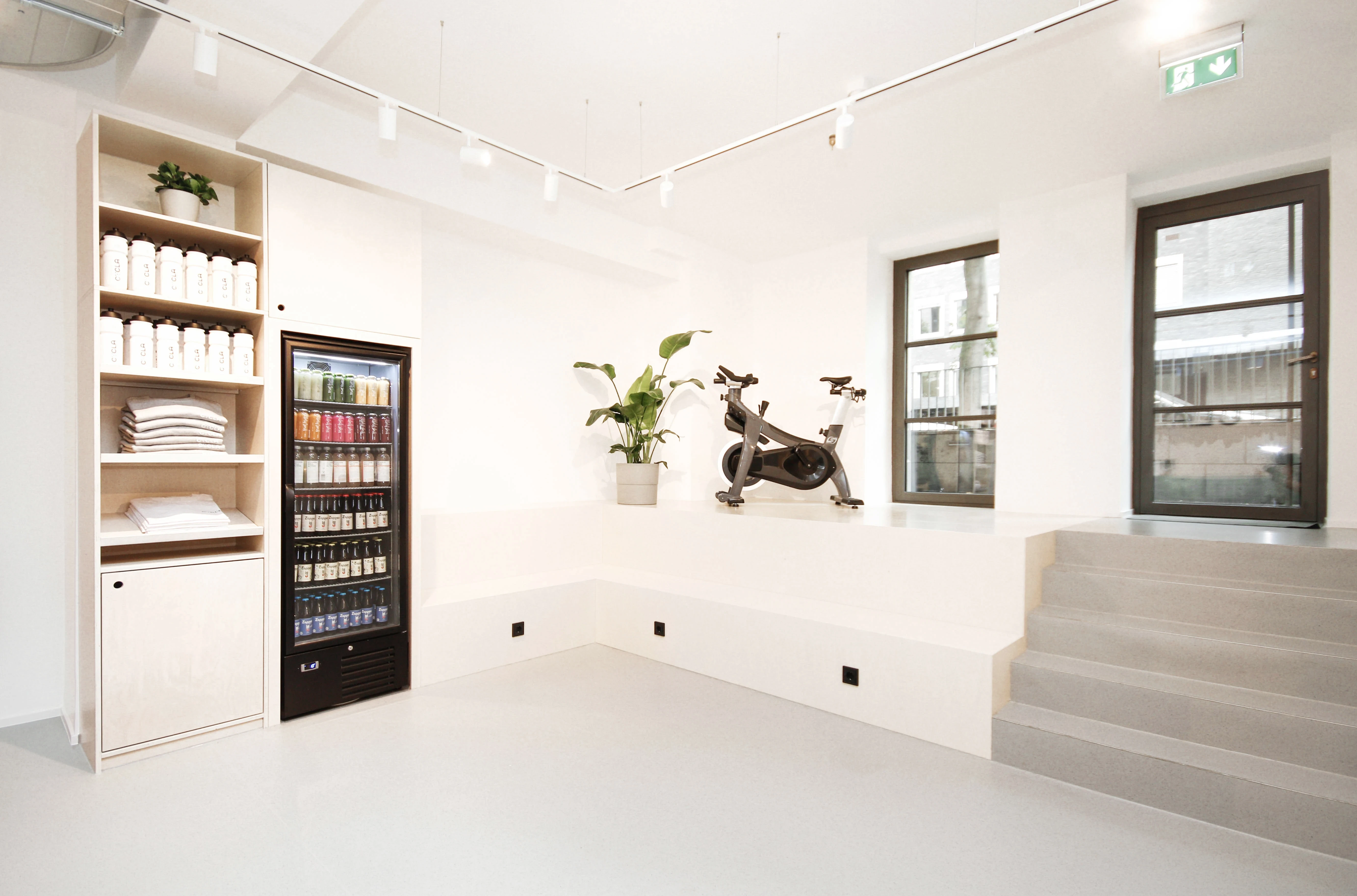 Shop corner at CYCLA with a wooden shelf of merchandise, a glass-front drinks fridge, a spin bike displayed on a white podium and steps leading to the window.