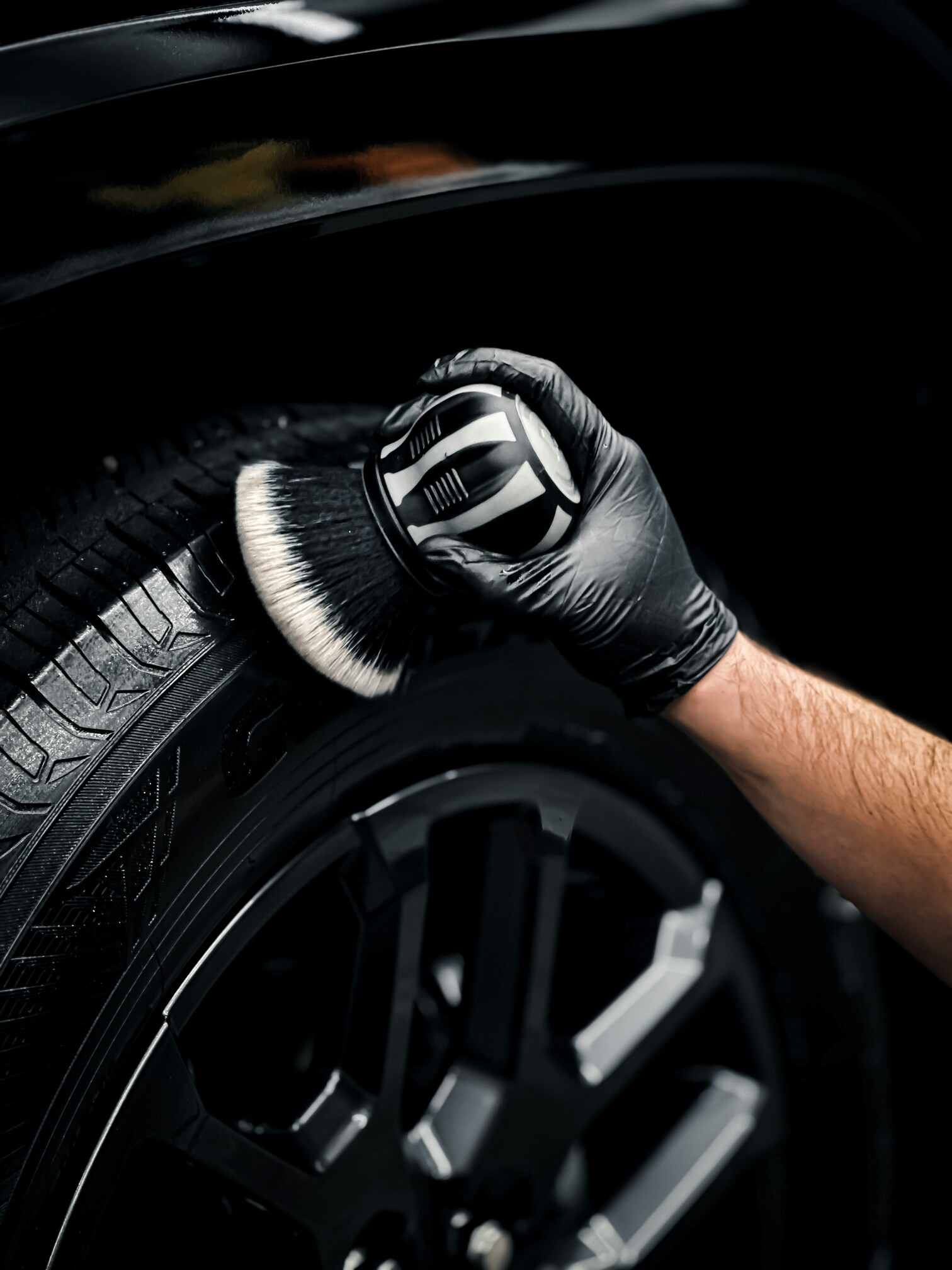 A person wearing a black glove uses a detailed brush to scrub the surface of a car tire, highlighting the tread pattern and rim design.
