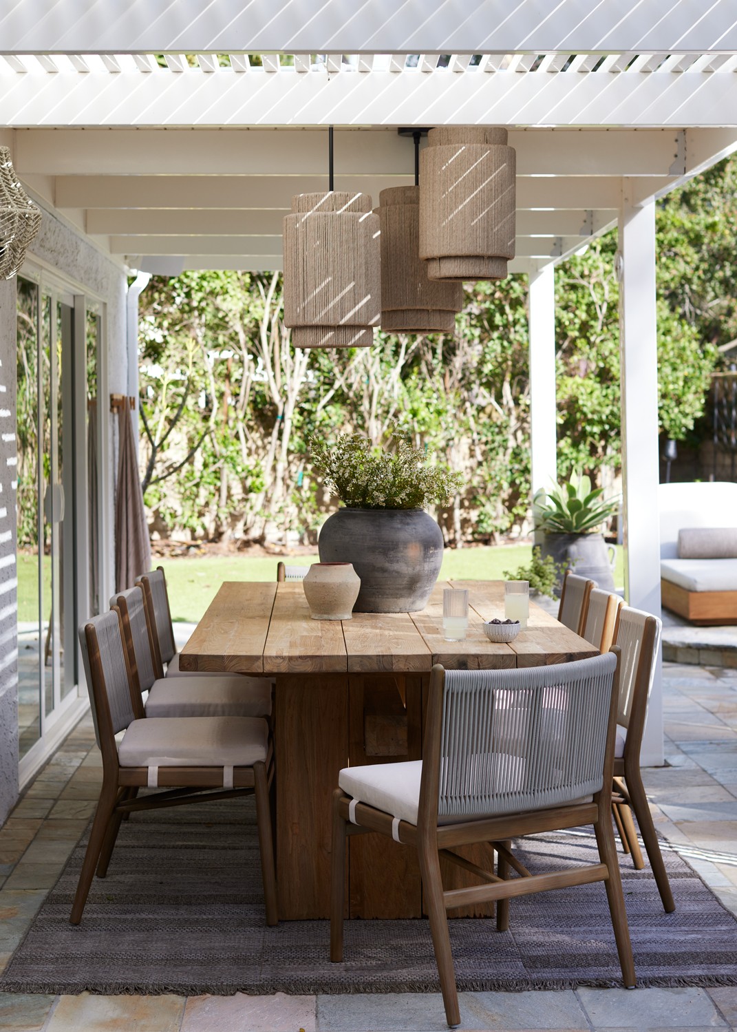 This landscaped outdoor retreat blends East Coast charm with Southern California sunshine, featuring a cozy patio, an eight-person wooden dining table, and rattan light fixtures for a warm, inviting ambiance