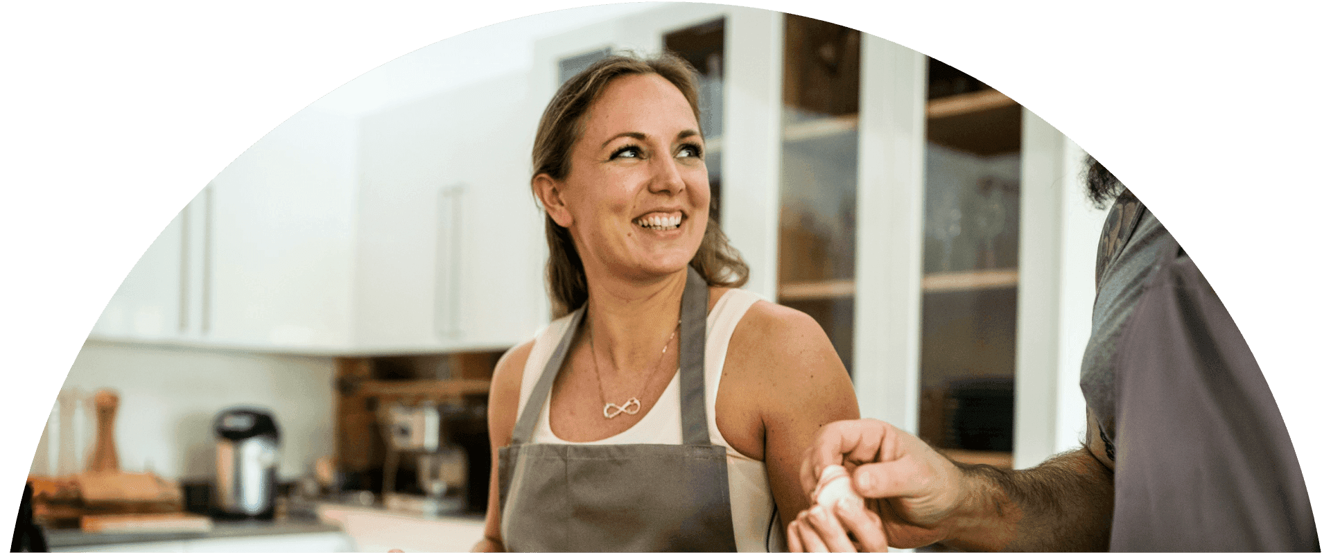 Woman in an apron smiles while cooking with another person in a bright home kitchen