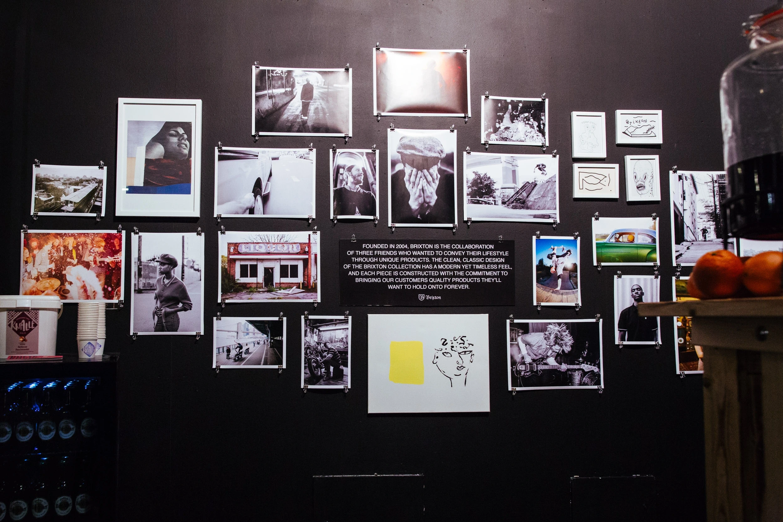 Black photo wall at the BRIXTON pop-up with clipped black-and-white lifestyle prints and a panel telling the brand story.