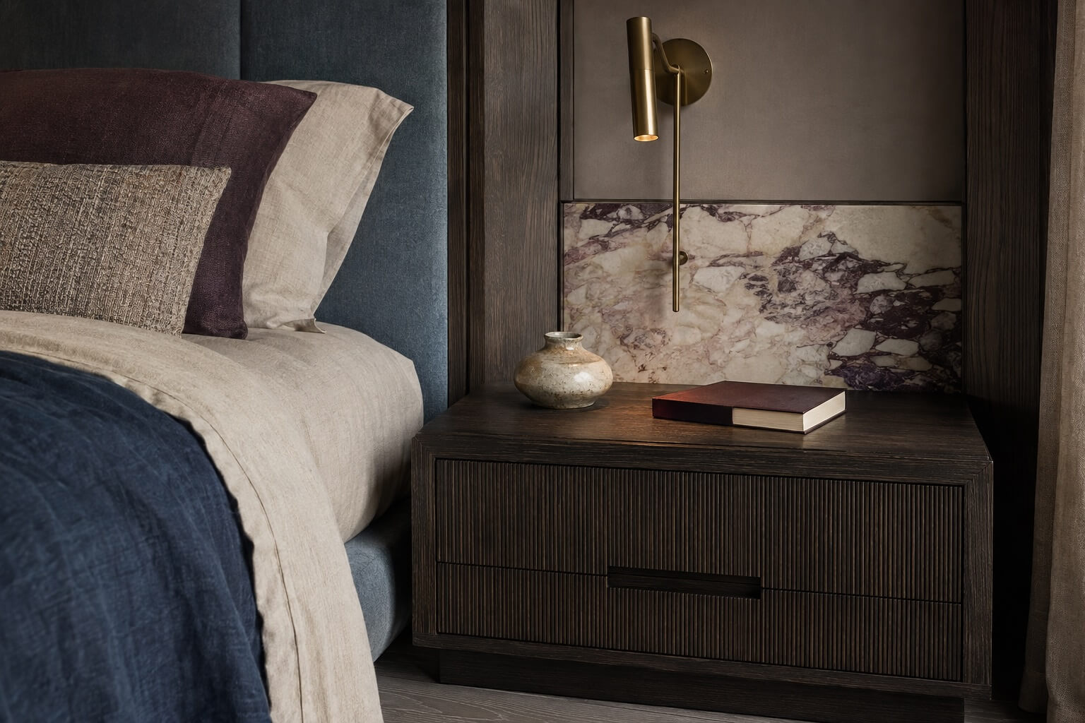 Luxury bedside detail with layered neutral bedding, a fluted dark wood nightstand, plum marble accent and brass reading light.