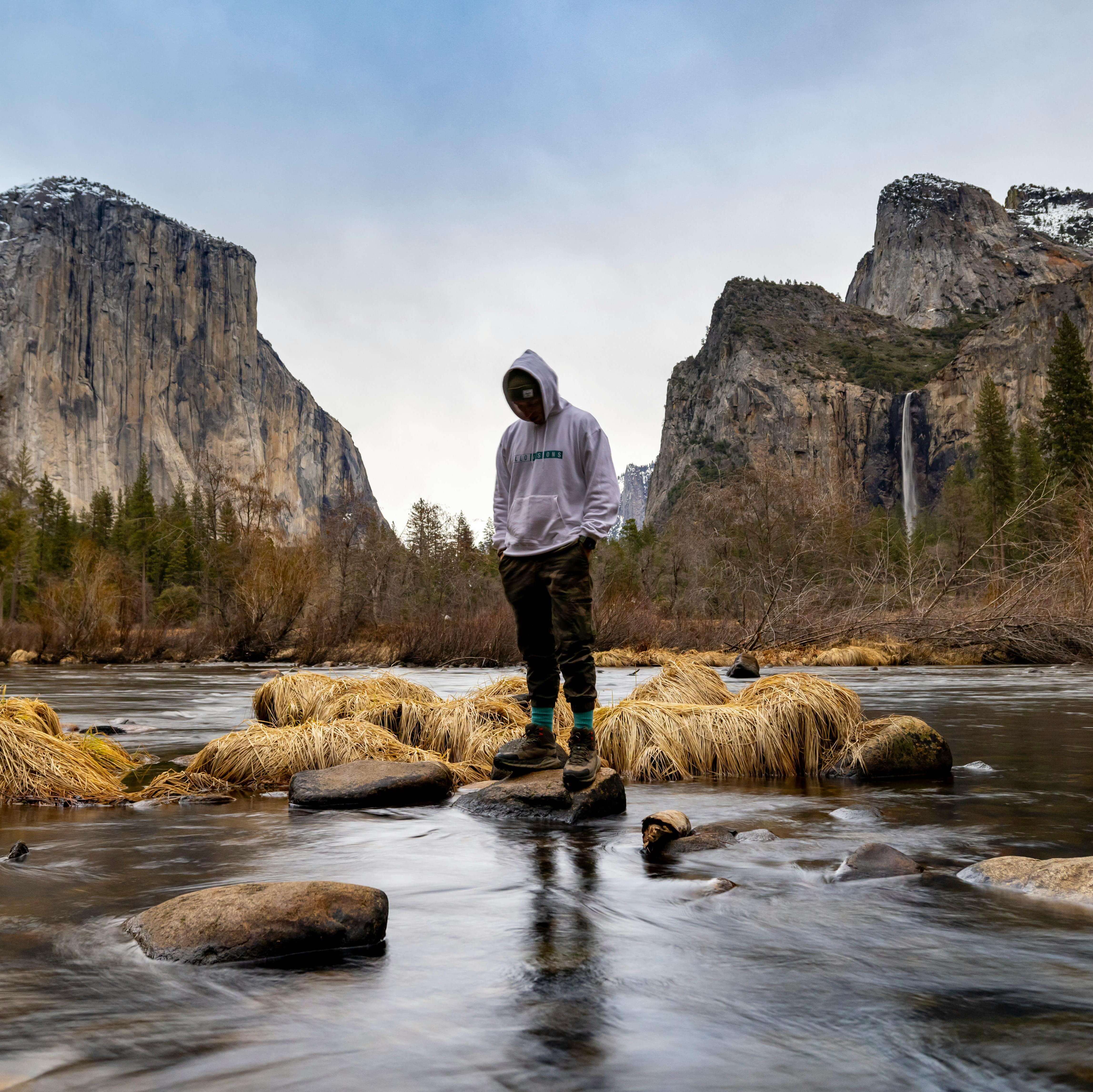 A person in a hoodie stands on rocks in a calm river, surrounded by autumn grass. Majestic cliffs and a waterfall are visible in the background. Peaceful atmosphere.