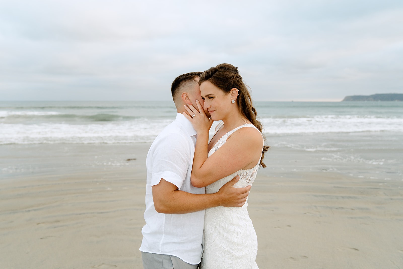 A San Diego couple embraces on the beach, smiling at each other with waves and clouds in the background.