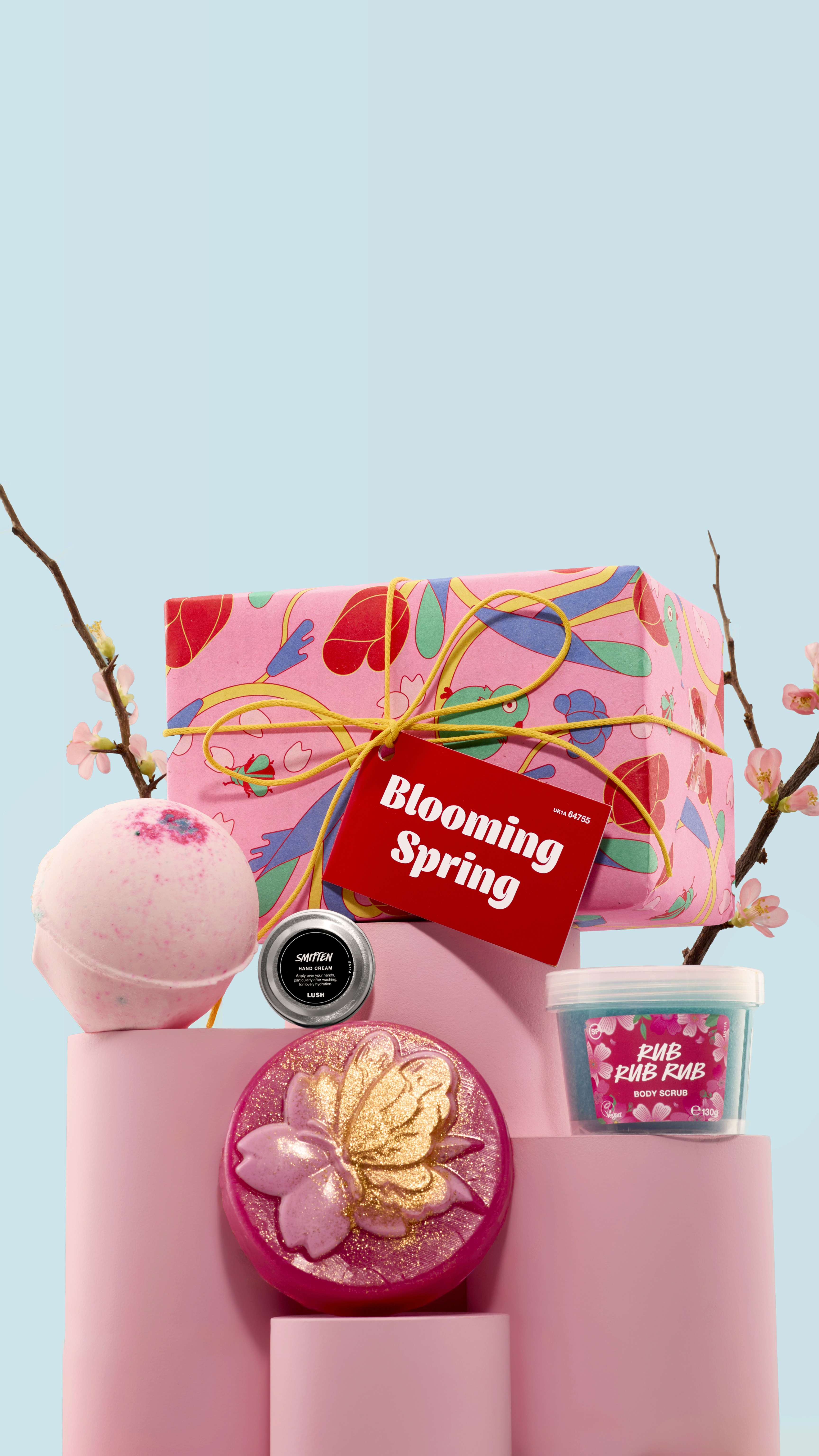 Lush “Blooming Spring” gift set with illustrated pink wrapping paper, yellow string, bath bombs, and small Lush containers displayed with cherry blossom branches on a blue background.