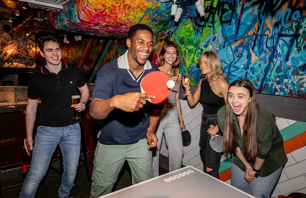 "Group of friends playing table tennis at a graffiti-walled venue, laughing and holding drinks, capturing an ordinary night out at a competitive socialising venue."