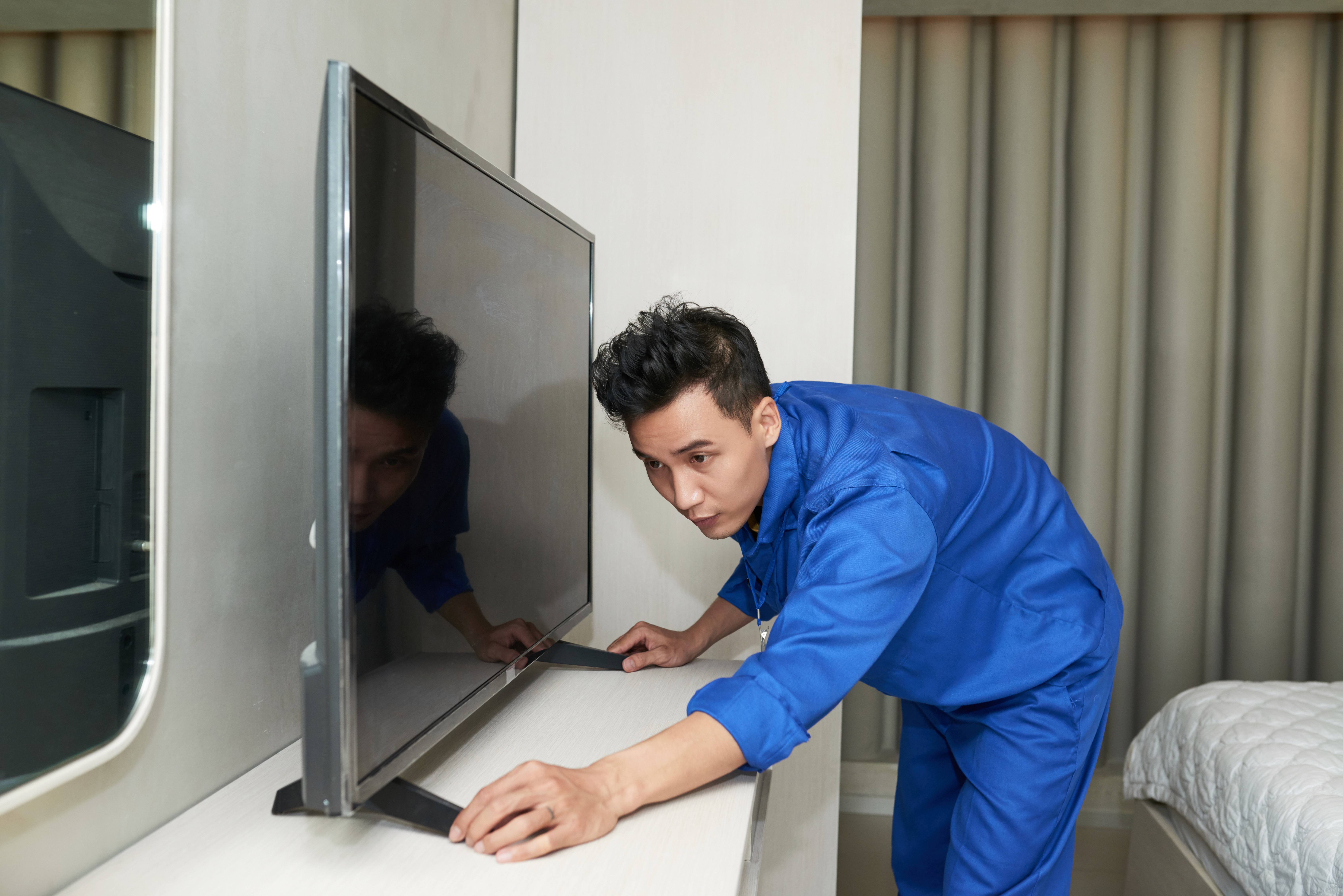 A man in blue overalls carefully adjusts and measures a flat-screen TV on a stand in a modern room. The setting is neat, with a bed visible in the background.