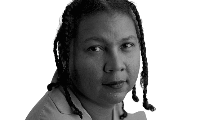 Black and white portrait of a woman with braided hair, looking directly at the camera with a calm, thoughtful expression. Her gaze is introspective, and the soft lighting enhances the contemplative mood. The simplicity of the background keeps the focus on her face, conveying a sense of depth and quiet strength.