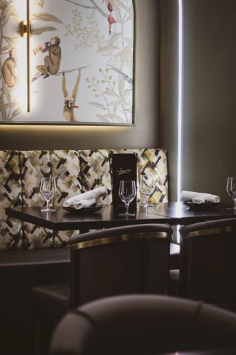 A restaurant booth with a dark wood table set for two, featuring patterned upholstery and a whimsical monkey-themed wall art.
