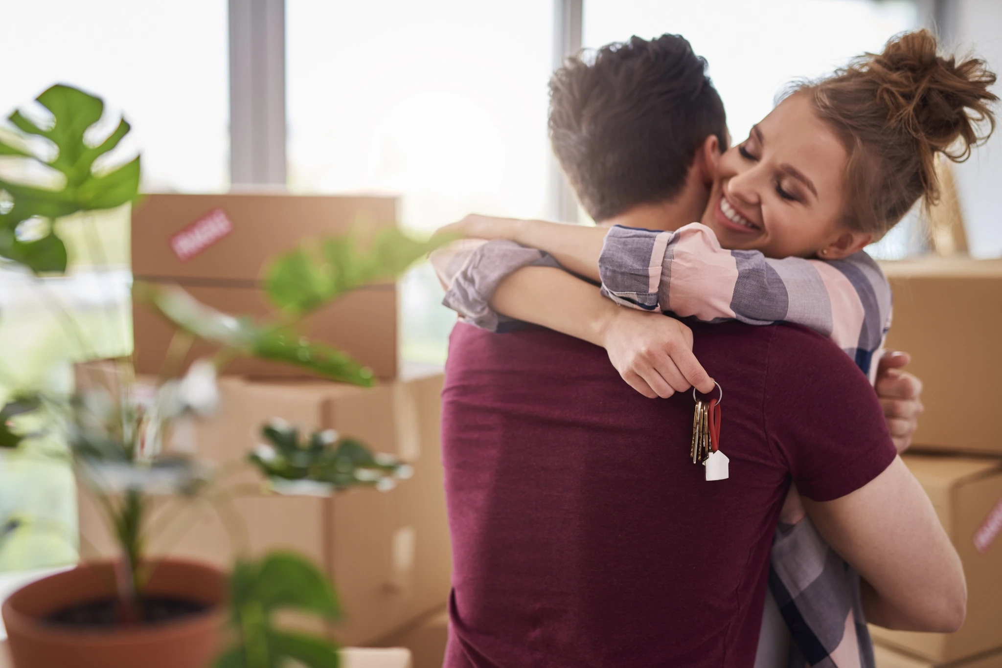 A woman smiles while being lifted by a man holding house keys during a move, surrounded by cardboard boxes and potted plants in a bright, sunlit room.