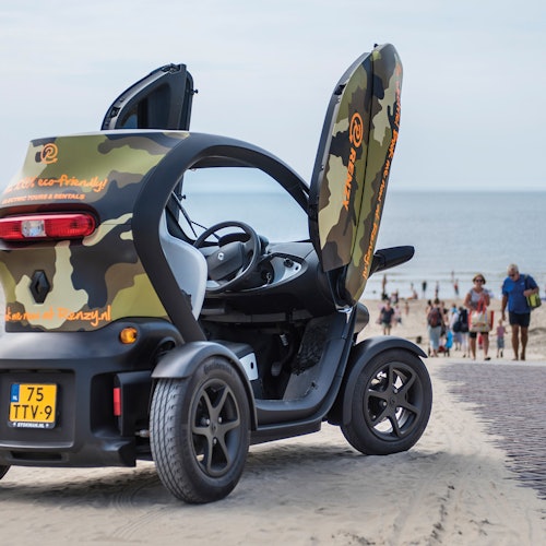 Sebuah kendaraan listrik kecil dengan cat kamuflase dan pintu terbuka diparkir di jalan berpasir dekat pantai yang ramai.