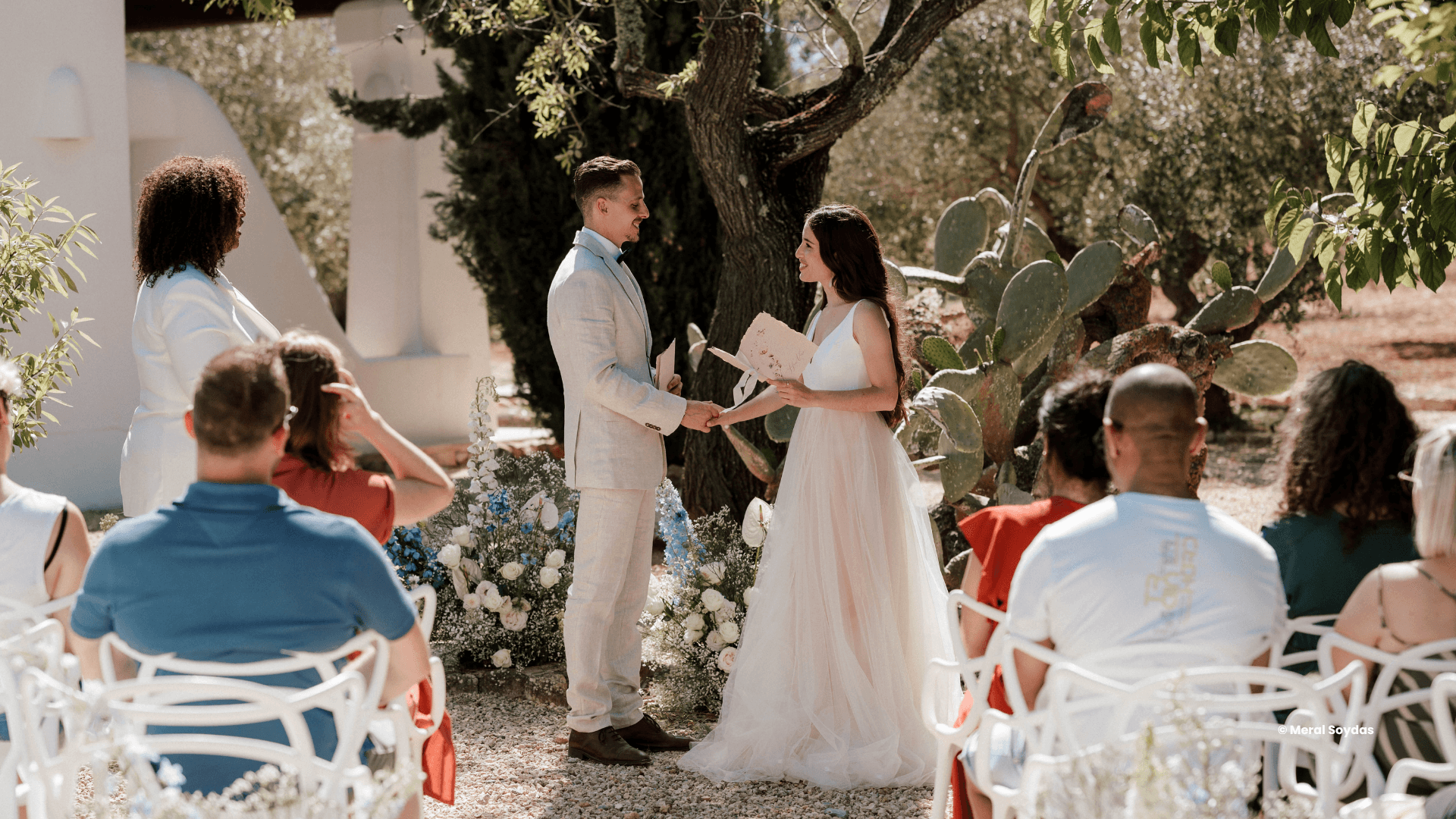 Een bruid en bruidegom staan tegenover elkaar met papieren in hun handen tijdens een buitenceremonie, geleid door een zelfstandig trouwambtenaar, terwijl gasten zitten en bloemen in de omgeving zijn opgesteld.