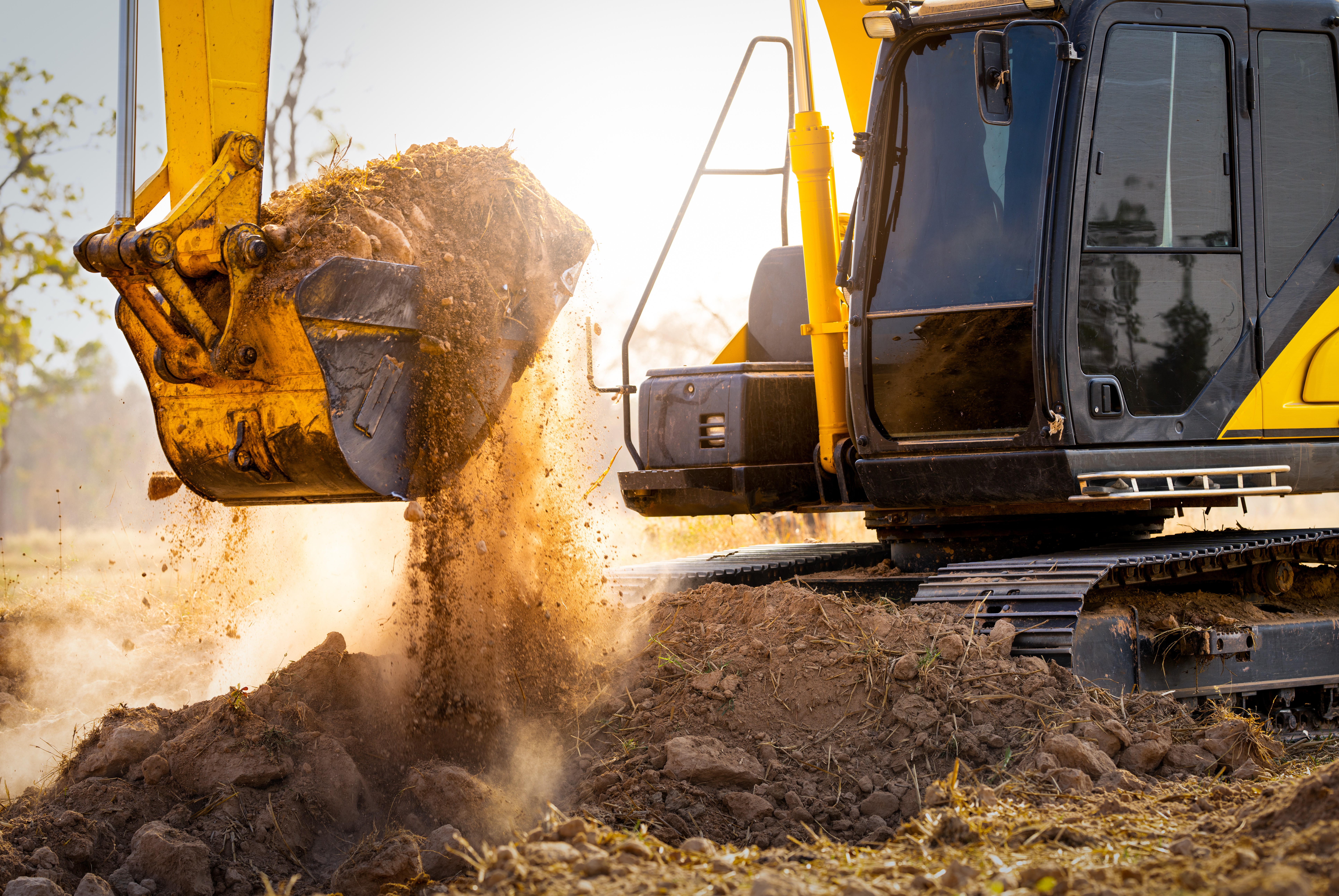 Close-up of a yellow excavator releasing soil from its bucket at a mining site, with dust particles illuminated by sunlight.