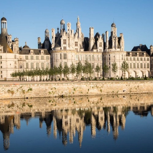 Large historic castle with numerous towers and spires reflected in a calm body of water on a clear day.