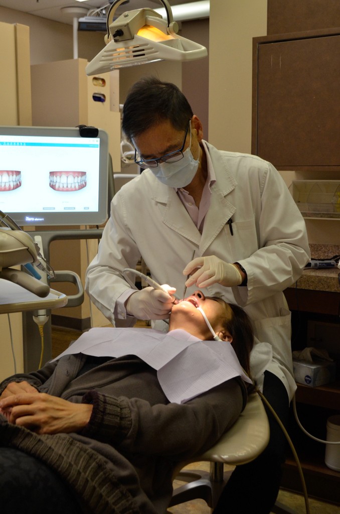 A dentist in a white coat and mask performing a dental procedure on a patient, with a computer screen displaying dental images in the background.