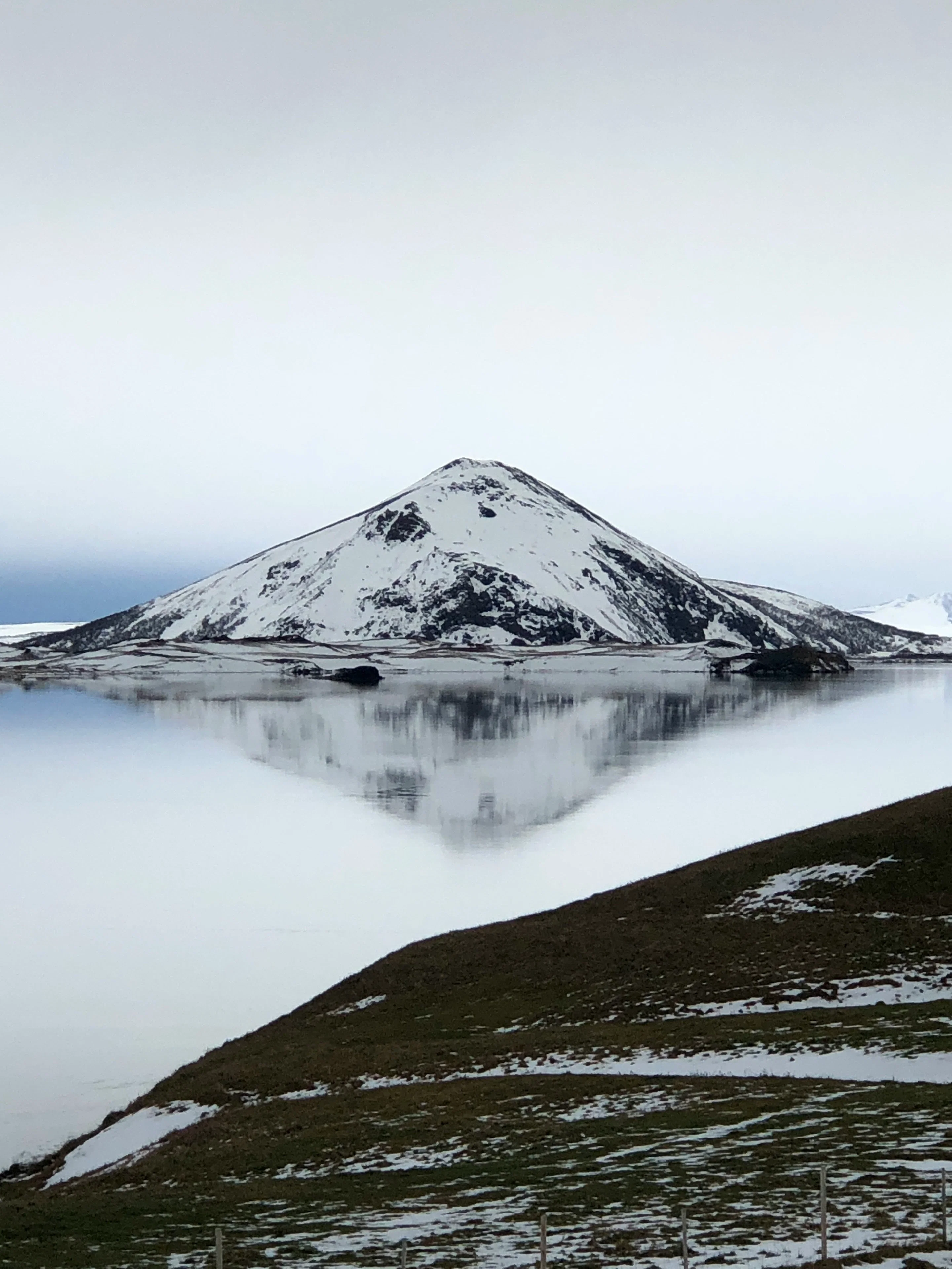 A snow-covered mountain perfectly reflected in the calm water of a lake with a snowy shoreline.