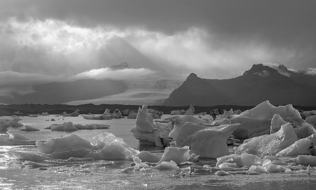 Icebergs floating in a glacial lagoon beneath dark clouds and distant mountains.