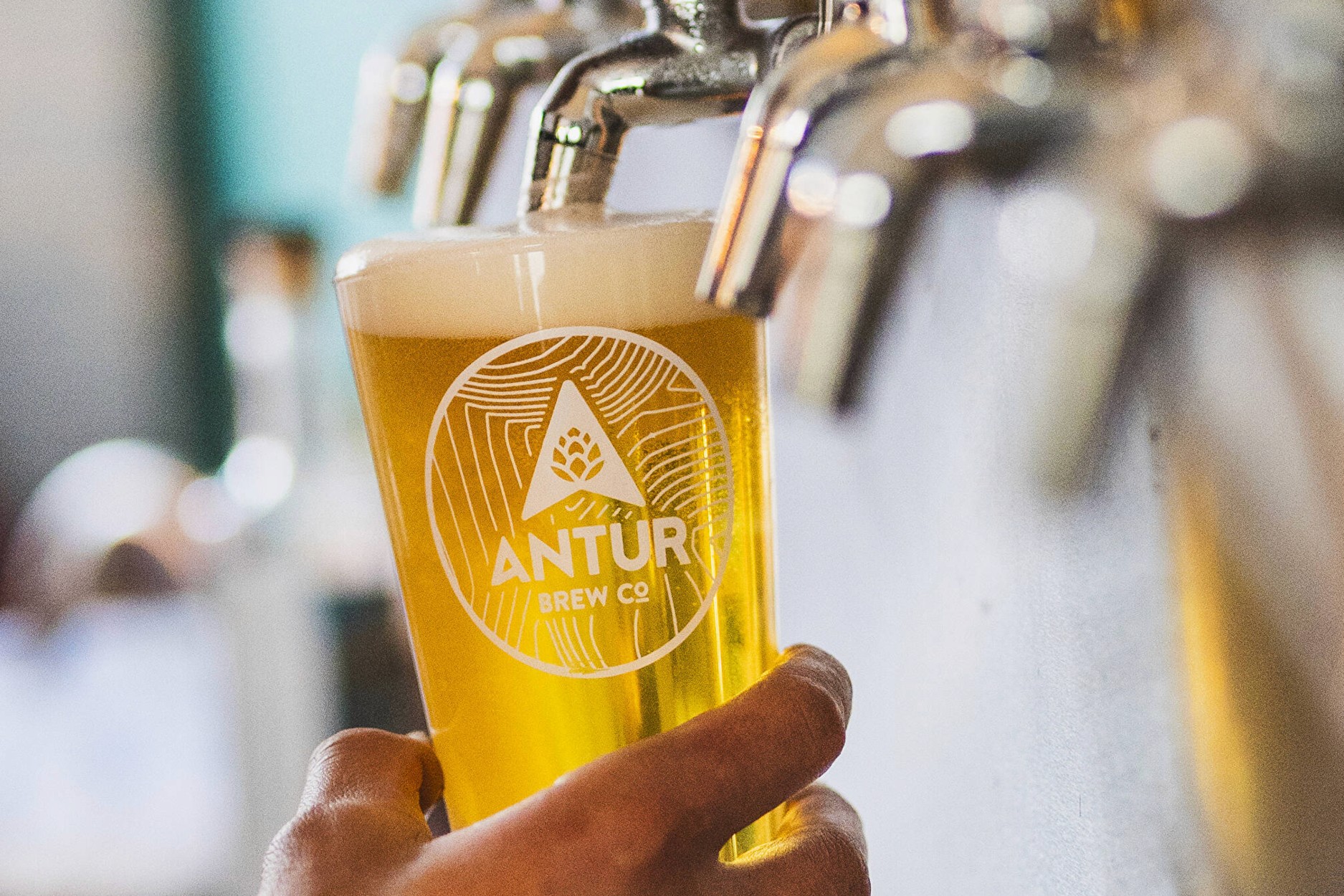 Hand pouring a pint of Antur Brew Co beer from a tap, with the Antur logo and topographic design printed on the glass.