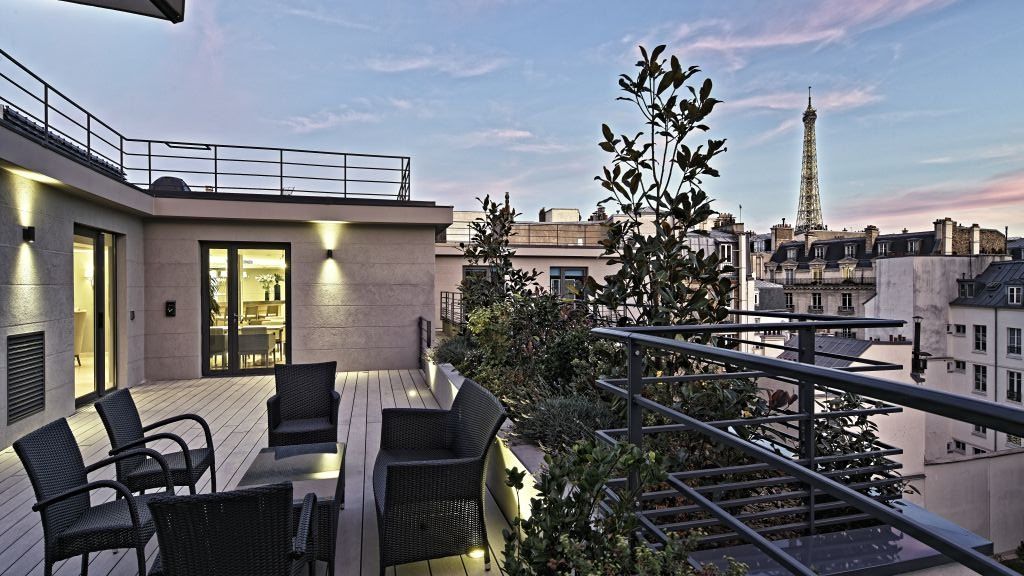 Rooftop terrace at dusk with wooden decking, black wicker furniture, planters with greenery, and iconic Eiffel Tower view in the background.