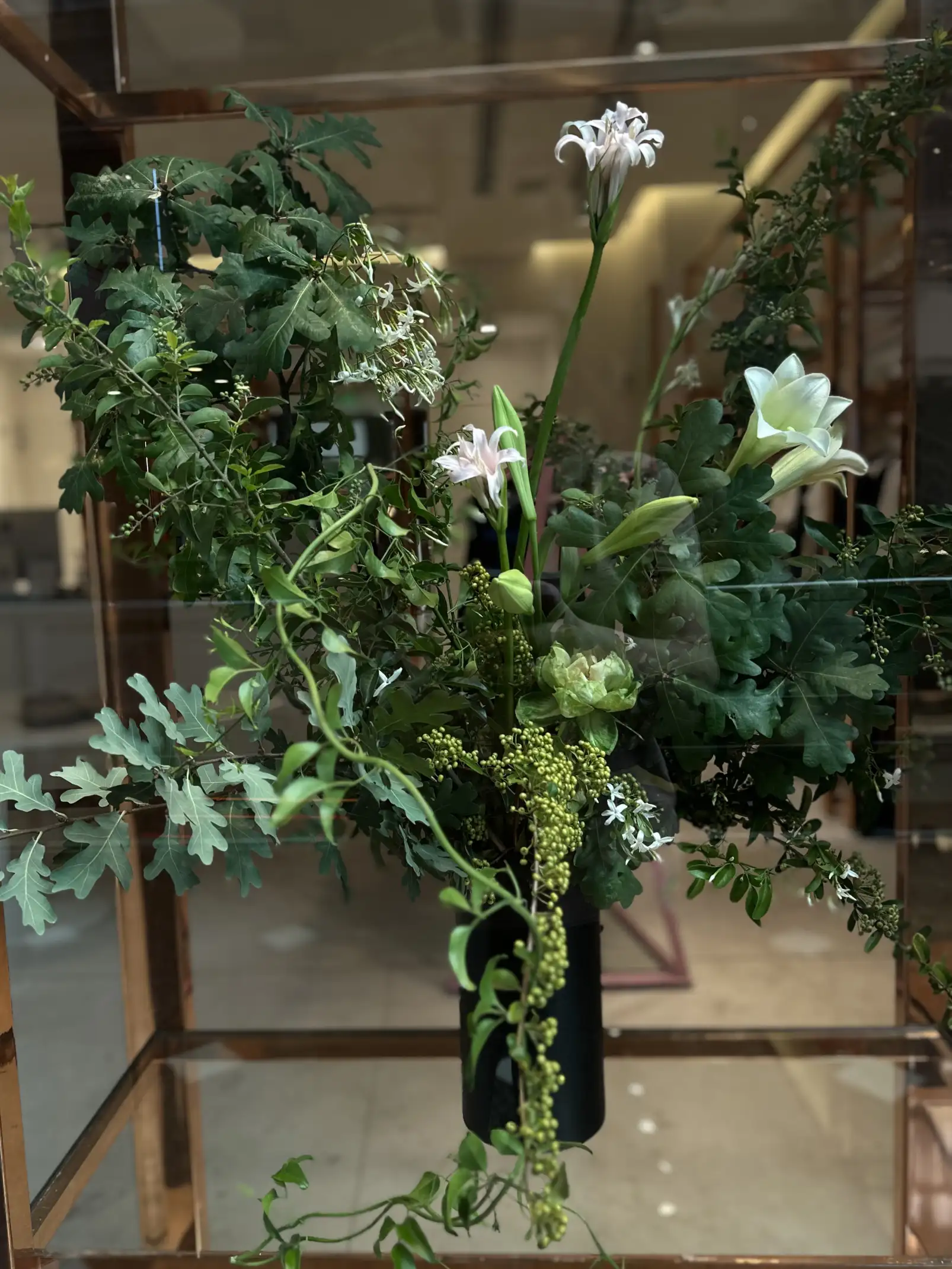 Composición floral contemporánea en jarrón negro con hojas y lirios blancos para decoración interior moderna.