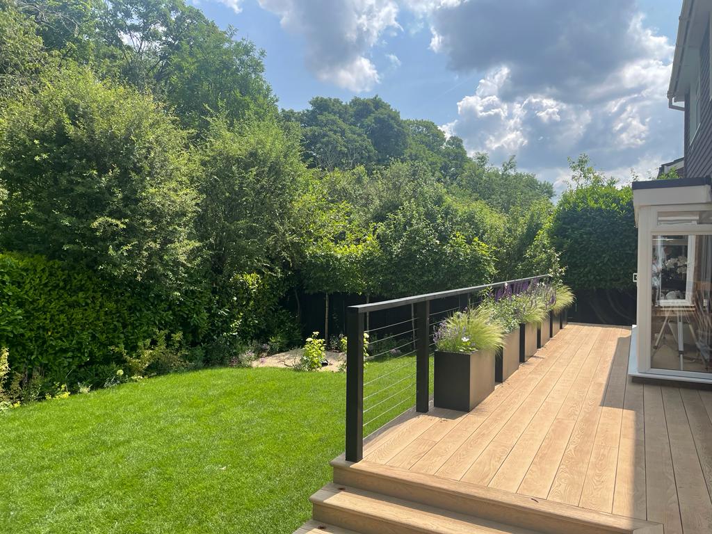A sunny backyard with green grass, trees, and a patio area, featuring a railing and clouds in the sky.