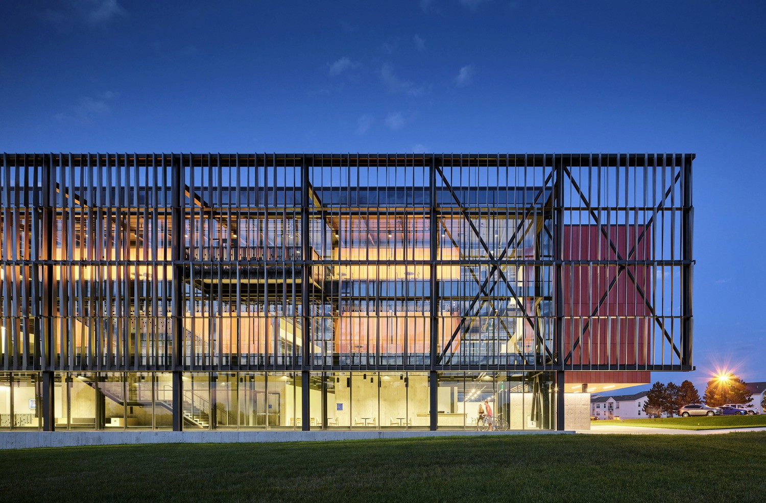 Modern three-story academic building at dusk with illuminated interior visible through floor-to-ceiling glass walls. The exterior features black structural beams in a grid pattern and a red accent wall. A grassy lawn extends from the building's base.