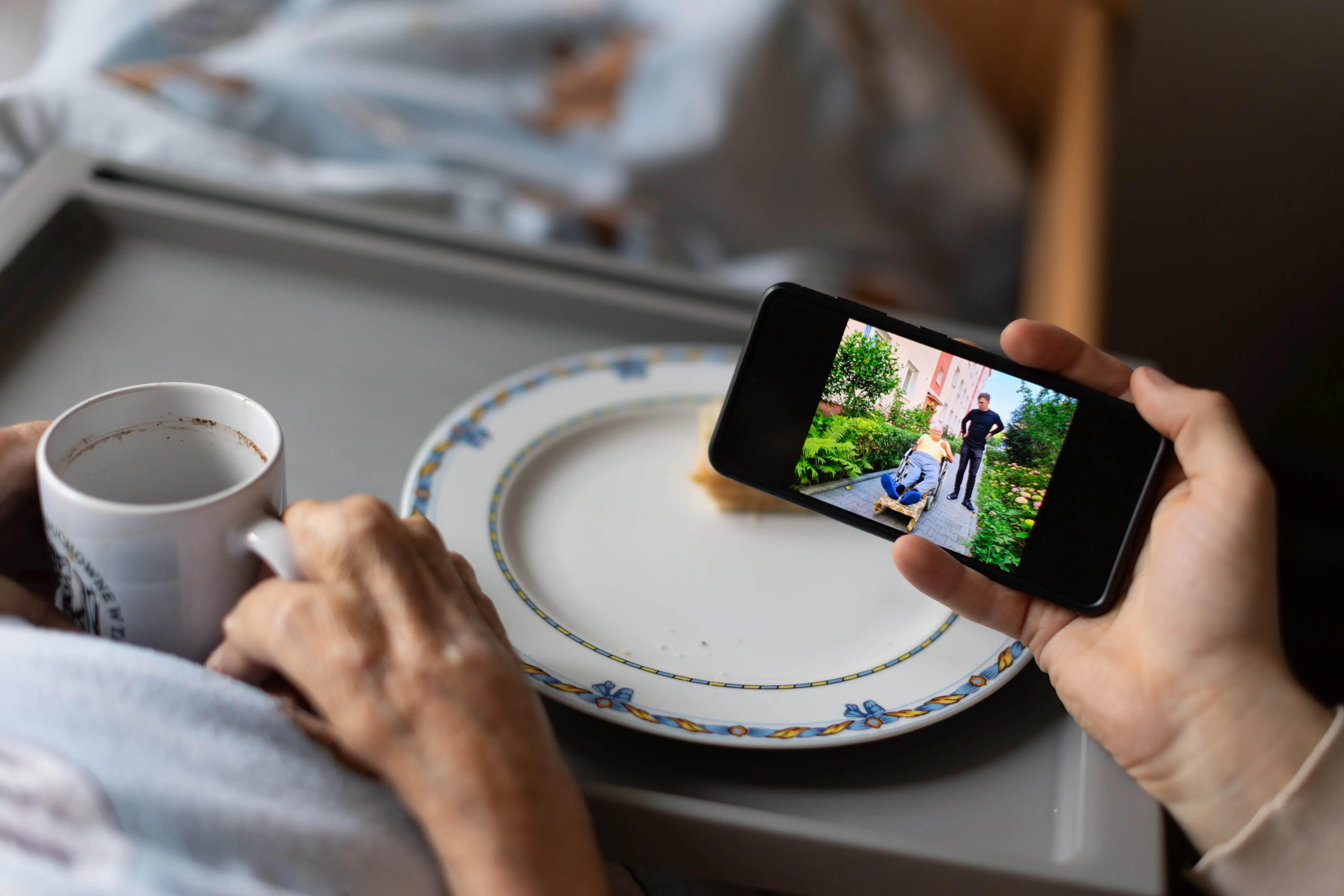 Ręka osoby trzymającej telefon, na którym widnieje zdjęcie. W tle znajduje się dłoń z kubkiem oparta na tacy z talerzem.