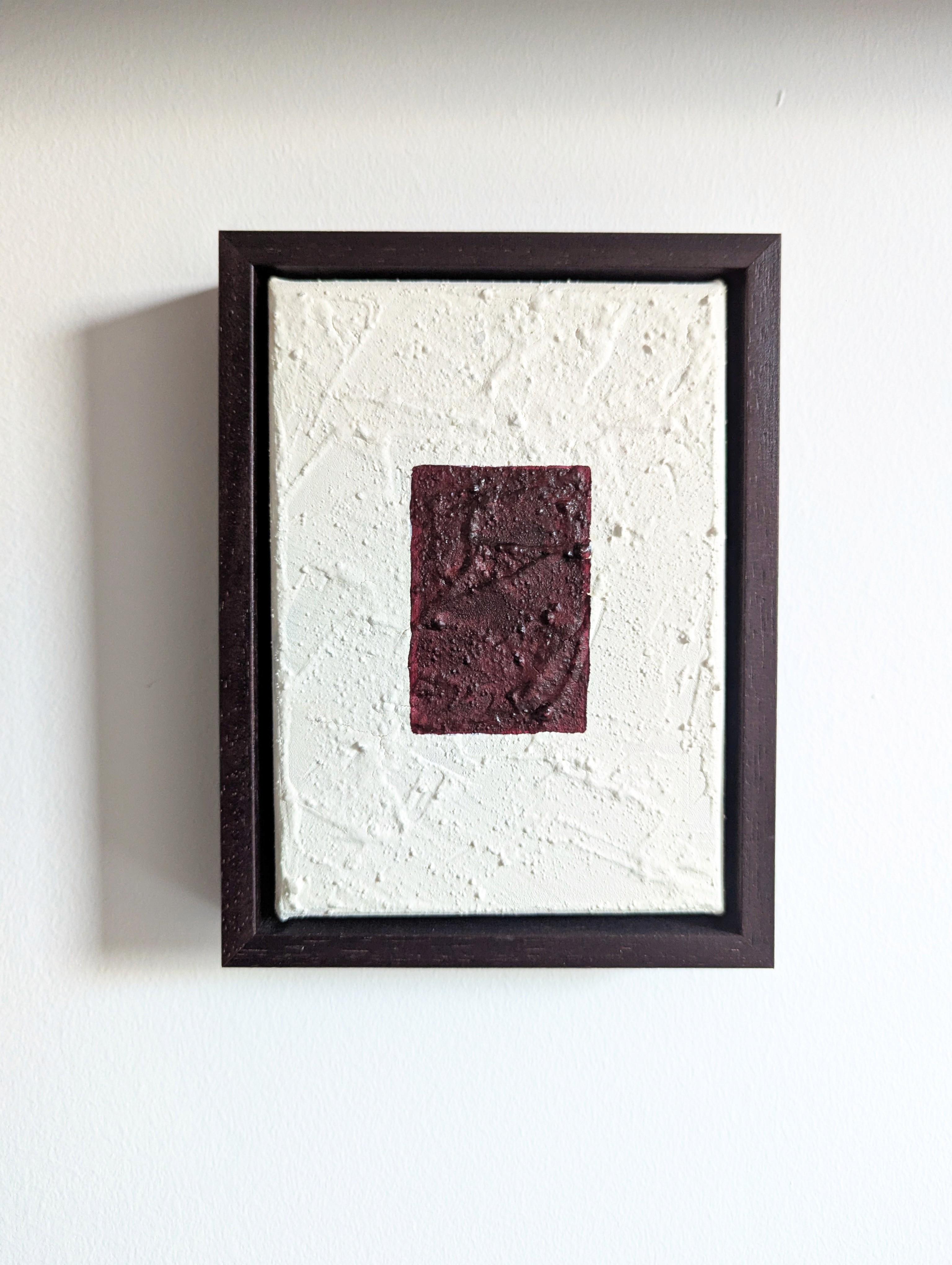 A framed art piece with a maroon design set against a white background, mounted on a wall.