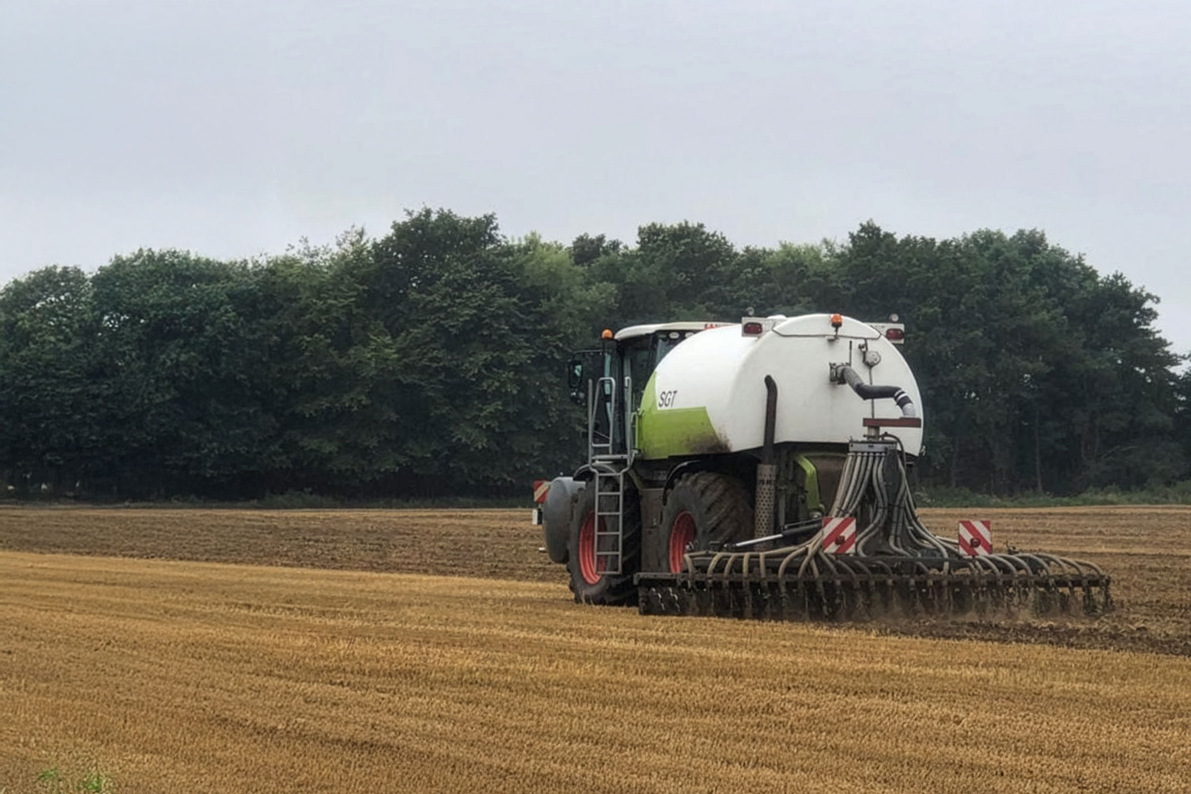 Claas tractor with white and green slurry tanker and trailing hose applicator working in stubble field with trees in background