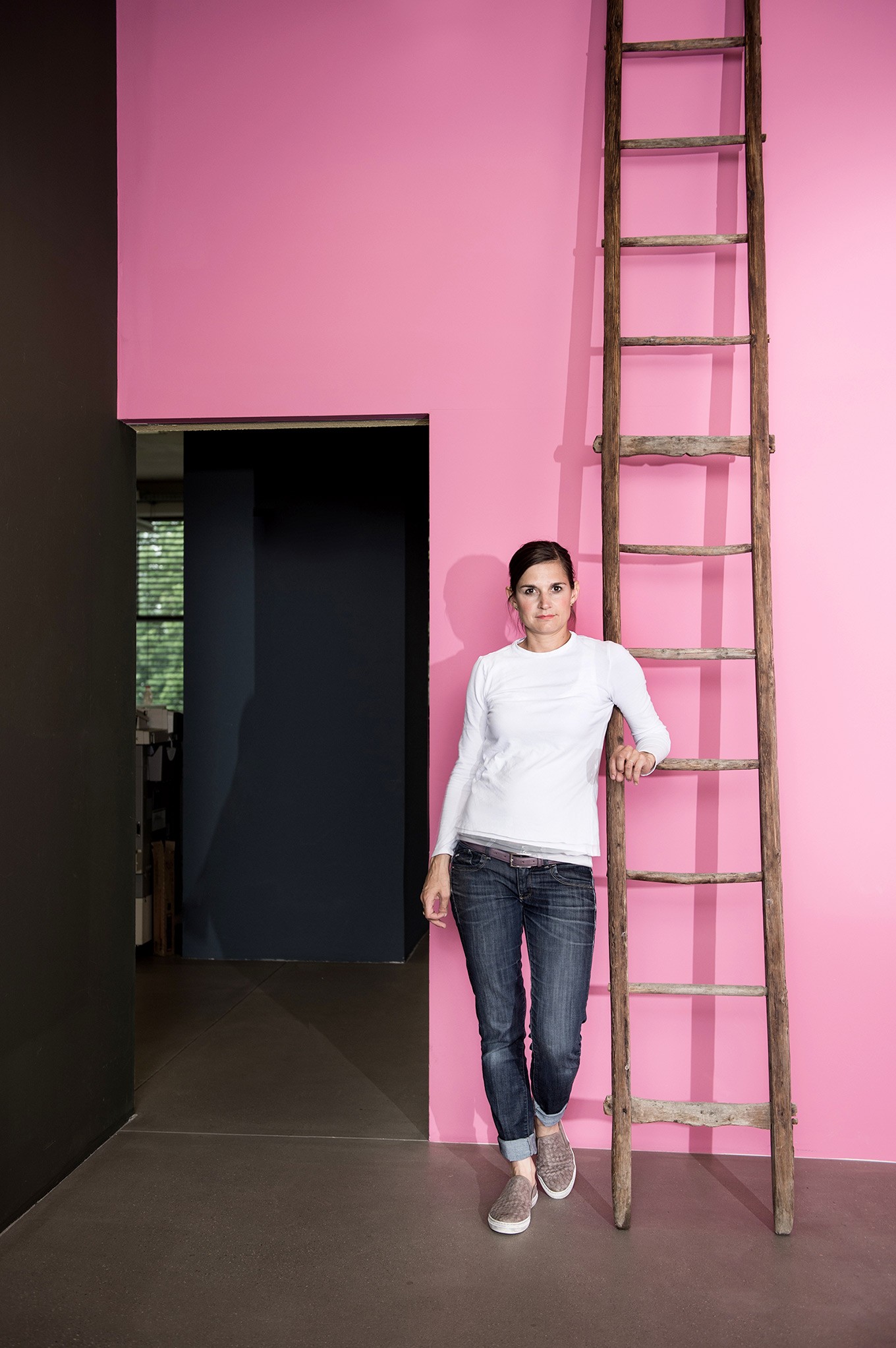 Fabienne Bühler in casual attire leans against a wooden ladder on a bold pink wall.