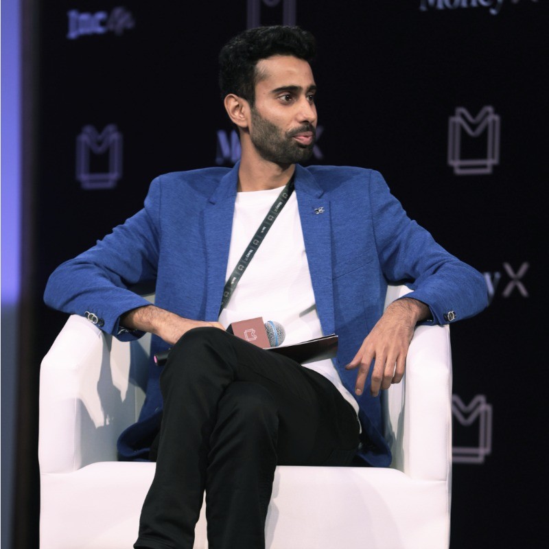 Man in a blue blazer sitting on a white chair on a stage, looking to his left.