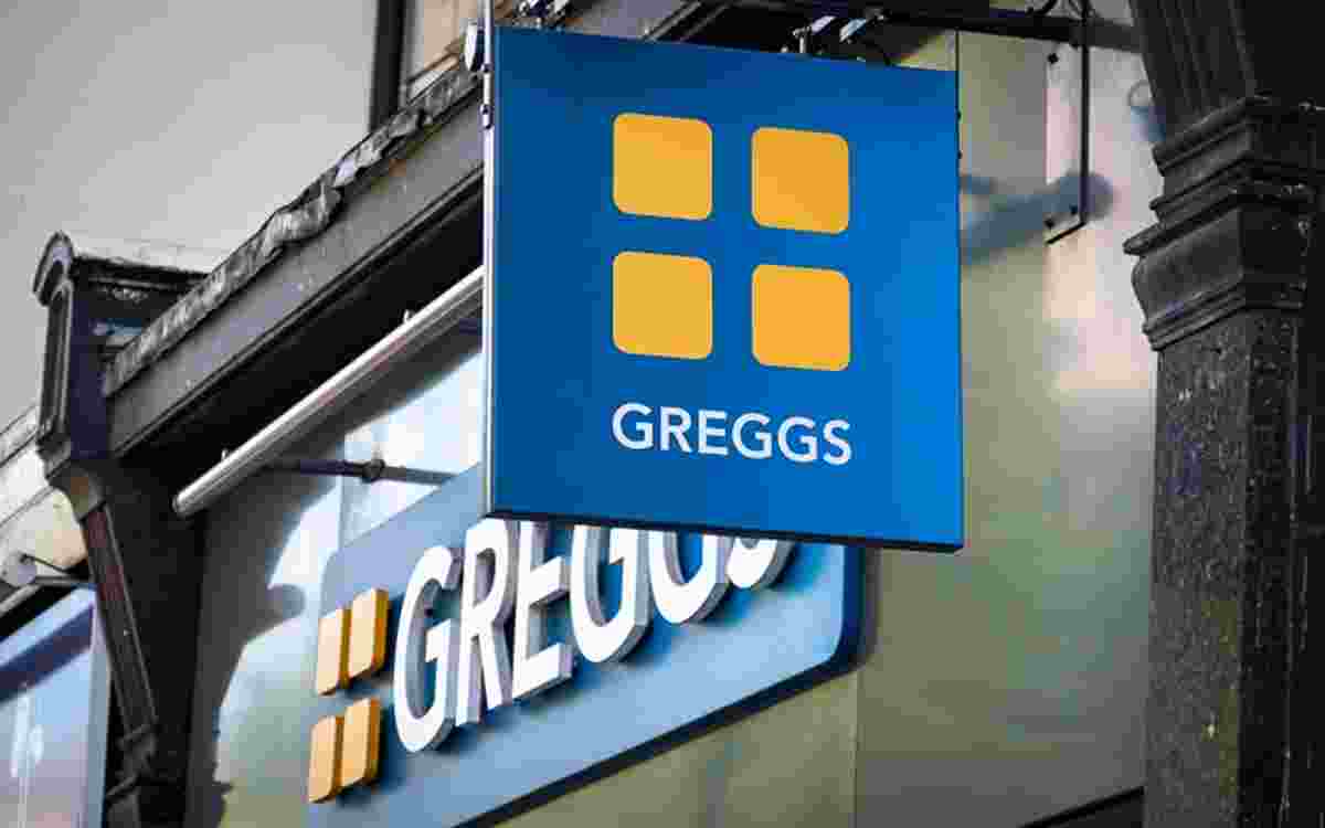 A blue Greggs shop sign hanging outside a building, featuring four yellow square shapes arranged in a grid above the word “GREGGS” in white capital letters. The sign is mounted on a metal bracket, with part of the shopfront and exterior architectural details visible around it, indicating a high‑street bakery location.