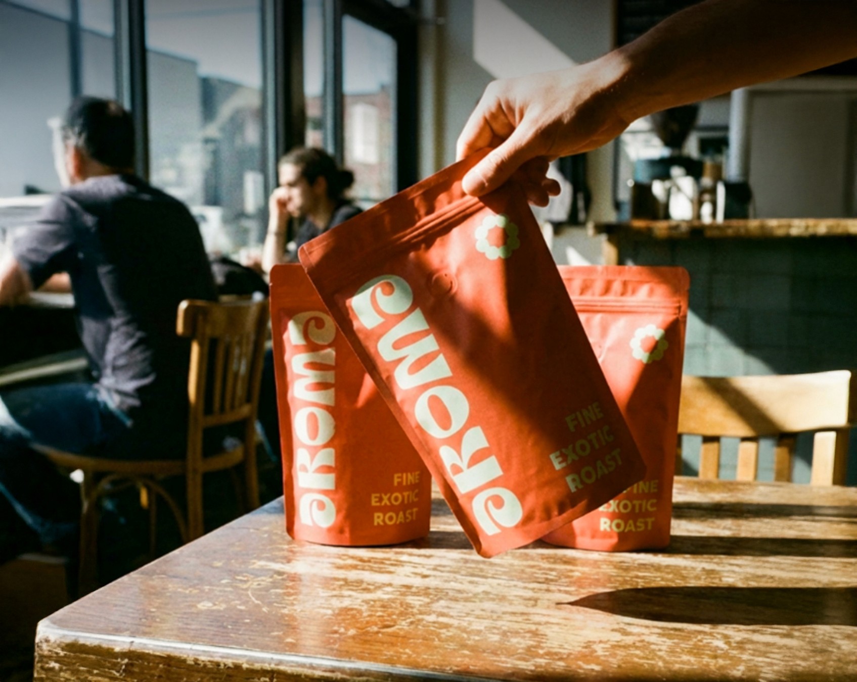A hand places a Aroma coffee bag labeled “Fine Exotic Roast” on a wooden café table, with two matching bags behind it and softly blurred people sitting near sunlit windows in the background.