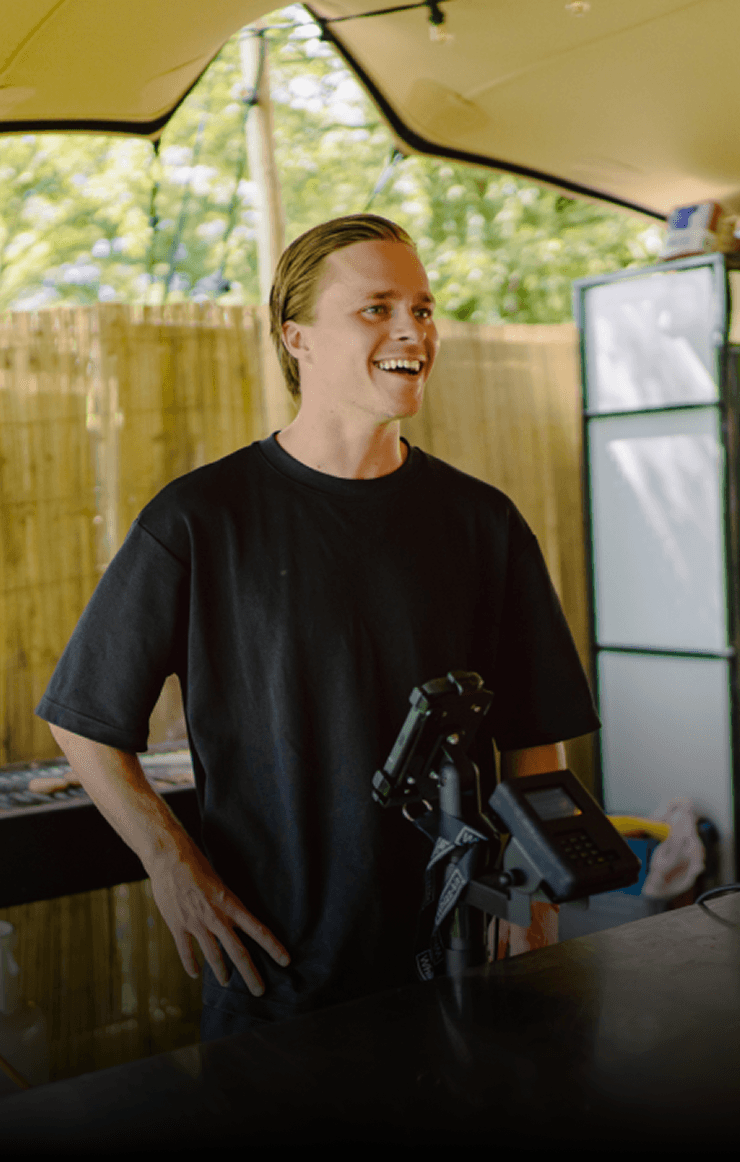 A smiling young man with crossed arms, wearing a plaid shirt and white t-shirt, poses against a dark background.