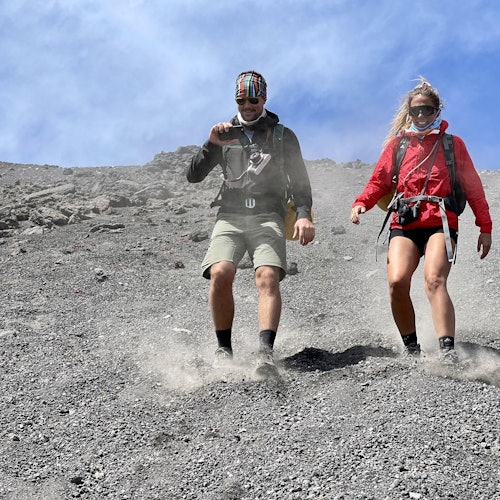 Duas pessoas vestindo equipamentos de caminhada descem uma encosta rochosa e empoeirada sob um céu azul claro.