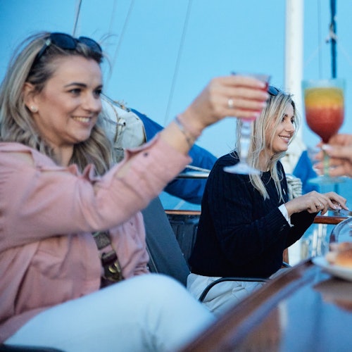 Two women seated on a boat, smiling and holding colorful drinks, toasting with another person.