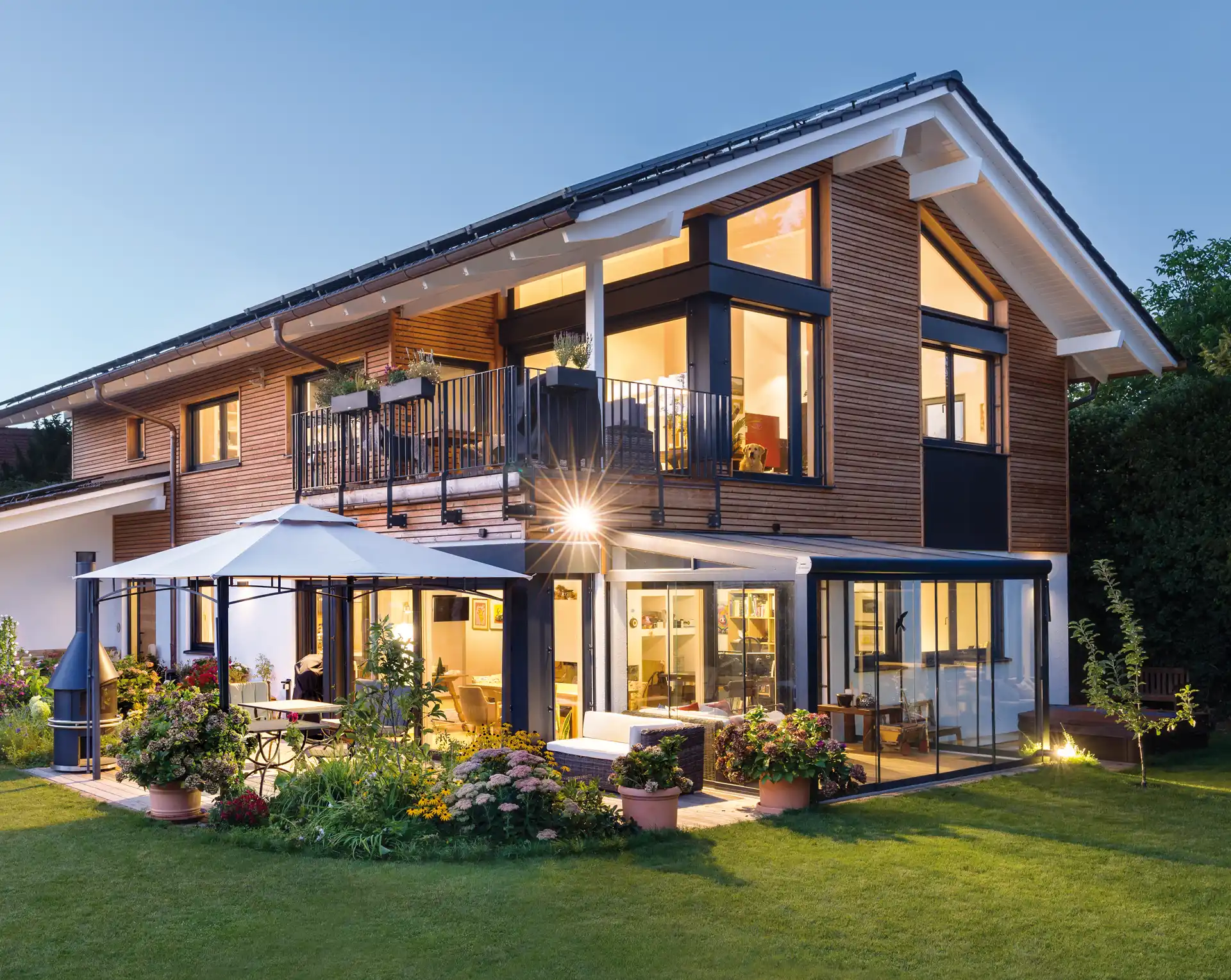 Geräumiges Holzhaus mit Veranda und Wintergarten, beleuchtet bei Dämmerung, mit Gartenmöbeln und blühendem Garten.