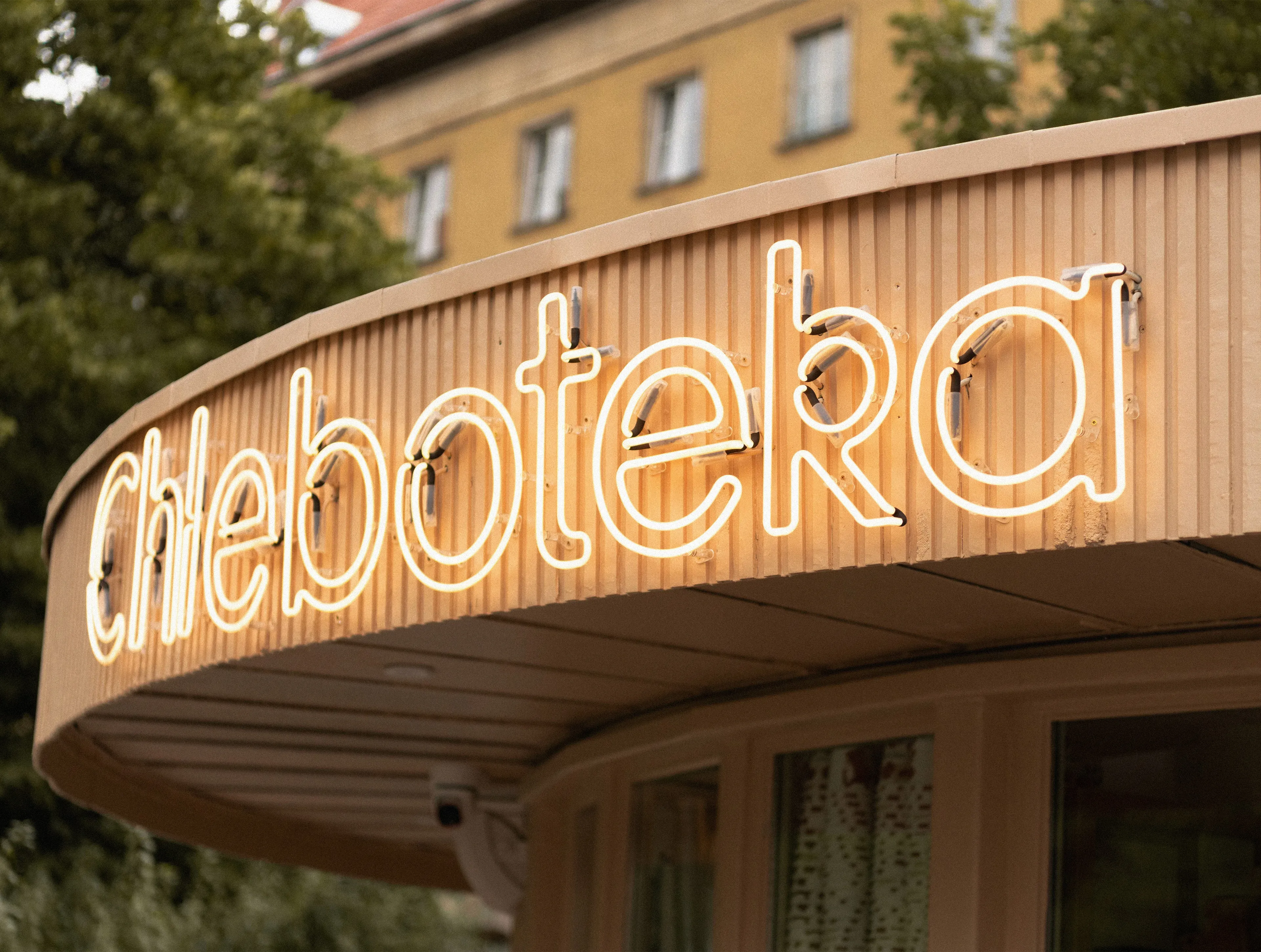 Chleboteka neon logotype sign on a ribbed beige pavilion facade, bakery signage and visual identity