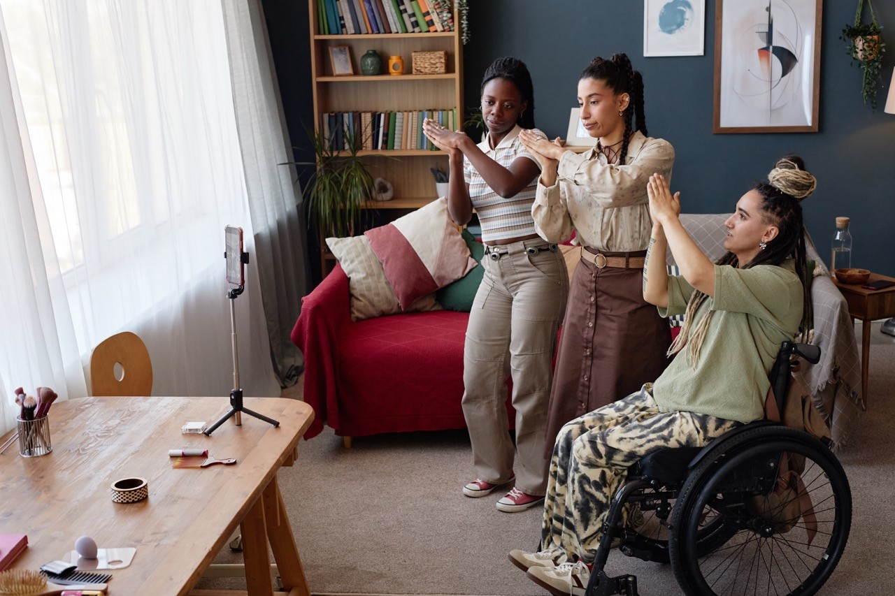Three women, one in a wheelchair, record a video together in a cozy living room. All are smiling and making hand gestures toward a smartphone on a tripod in front of them.