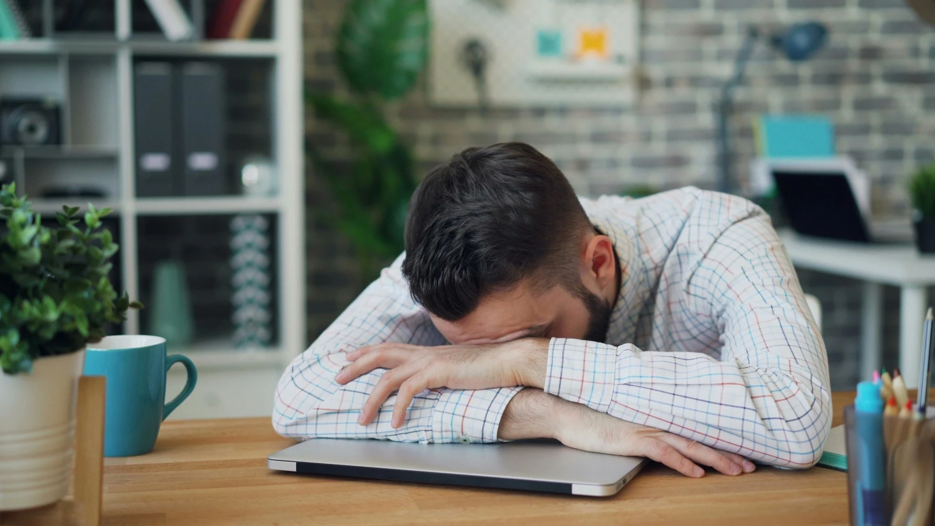 A young professional (early-career new hire) in a patterned shirt is slumped in exhaustion over a closed laptop at an office desk, illustrating the critical problem of 'new hire burnout' discussed in the C2 Essentials article.