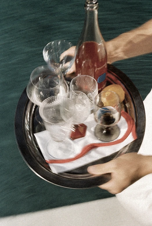 A close-up of a tray being carried by hand, featuring a bottle of rosé, elegant glassware, and citrus garnishes — evoking effortless luxury and summer indulgence.  