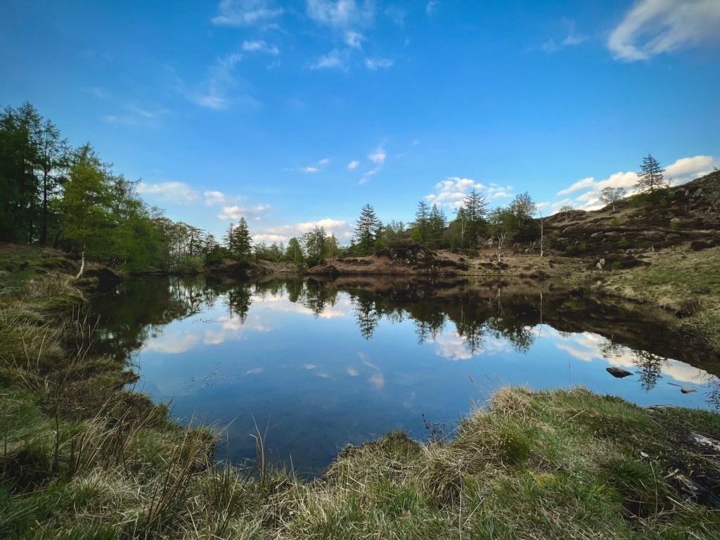 A wide-angle shot of the reservoir (tarn). The still water reflects the trees behind and the blue sky above.