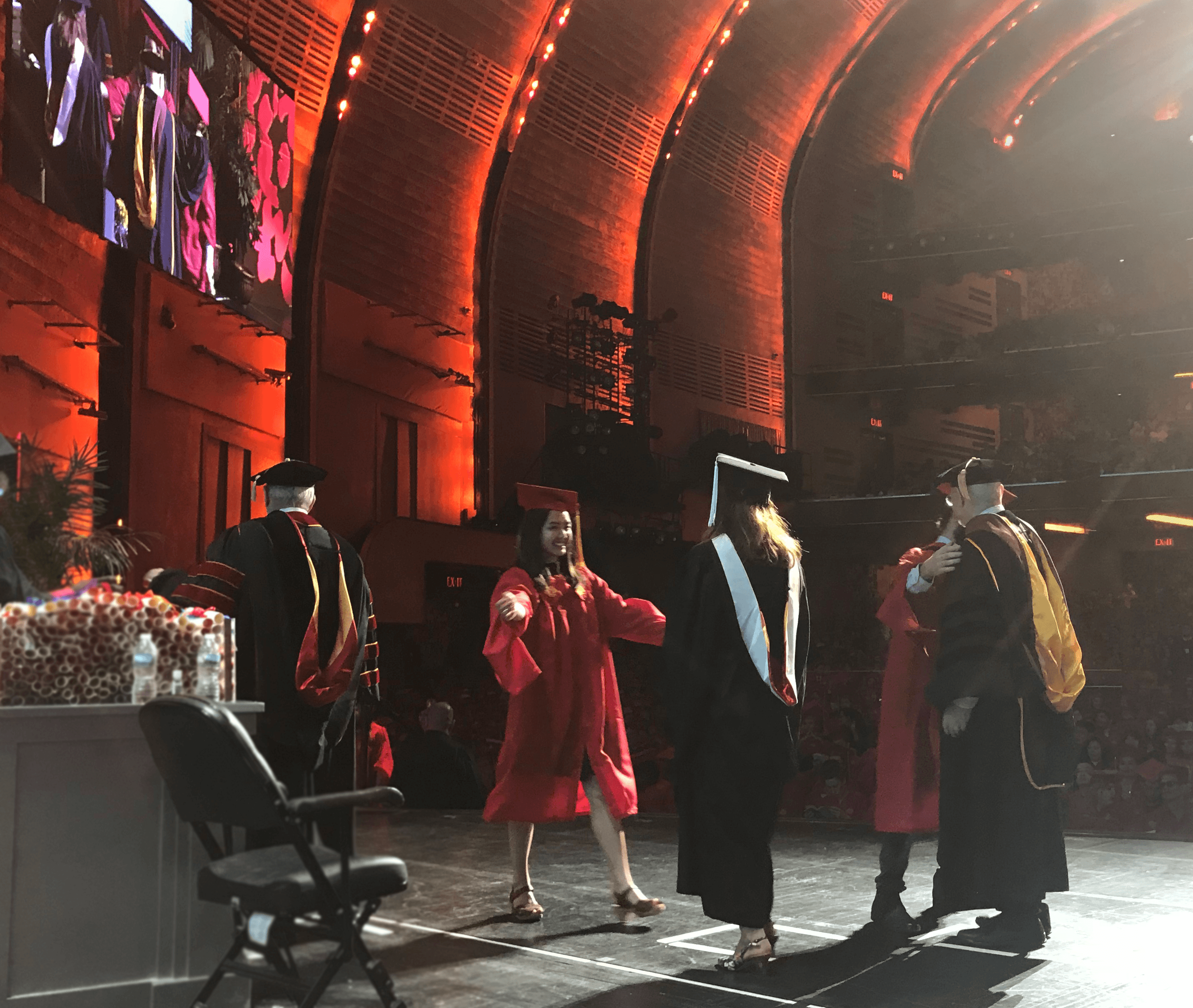A photo of a group of students and teacher on a stage at the graduation ceremony. They are wearing black or red robes and hats.